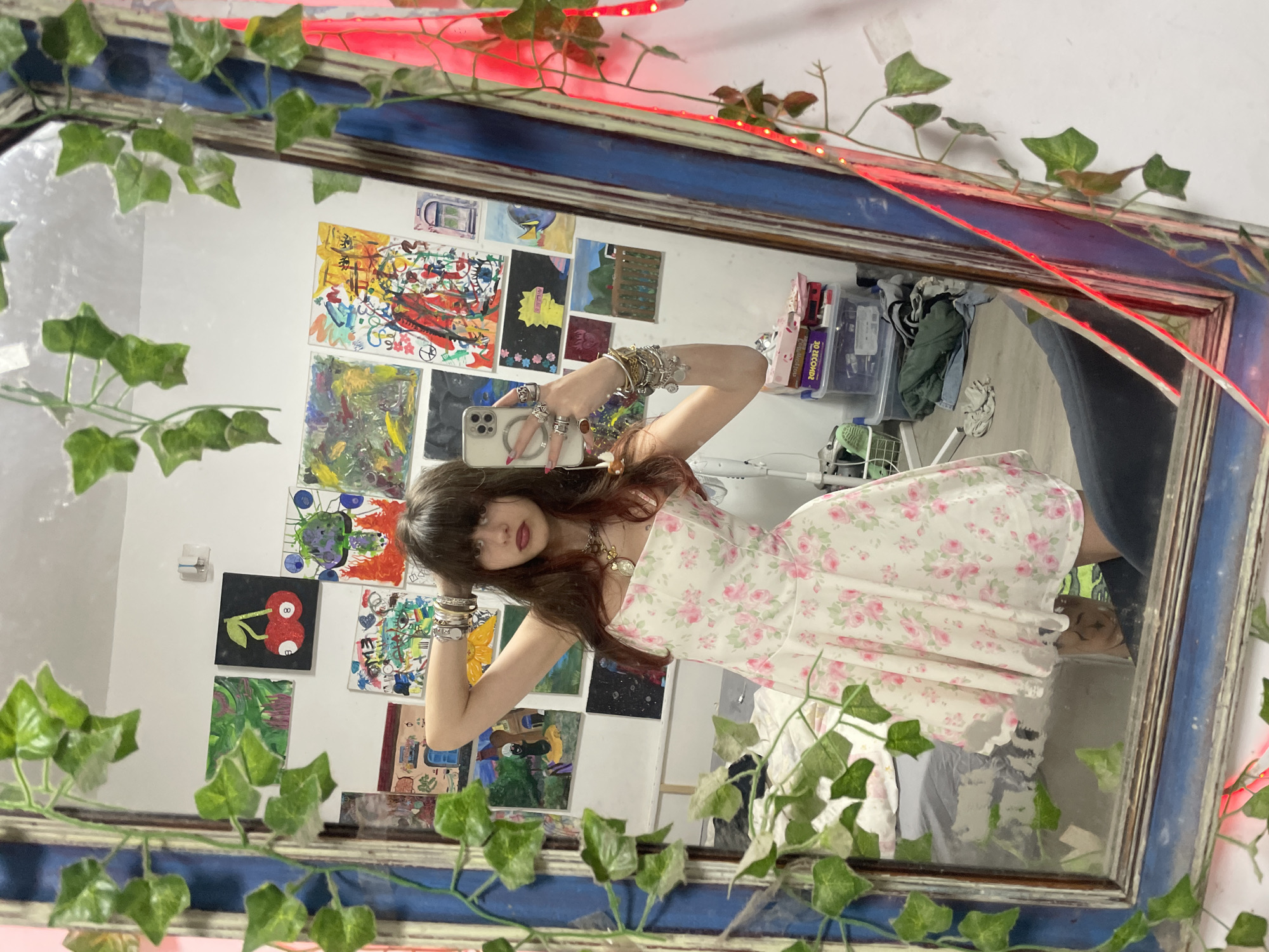 The image depicts a person with long brown hair and bangs, wearing a white floral dress and numerous bracelets, taking a selfie in a mirror. The mirror is decorated with artificial ivy and a red string light. The background visible in the mirror includes a white wall covered in various colorful paintings and drawings. A cluttered storage area with bins and clothing is also partially visible. The location is presumably indoors but there is no information in the image that suggests it is in Raanana, Israel. There are no actions or interactions, just a portrait.