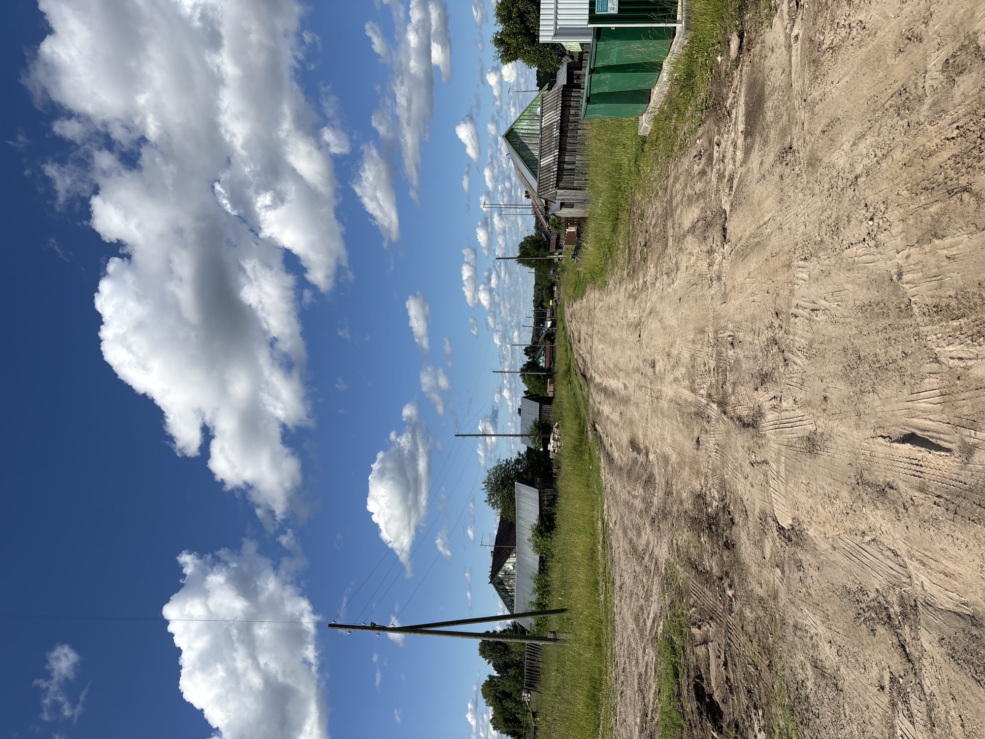 This is a landscape image captured in a rural village setting, likely in Kurgan, Russia. The photograph is oriented vertically, presenting a view down a dirt road that runs along the right side of the frame. The road is rough, with visible tire tracks and patches of loose soil and gravel, suggesting it's a frequently used path.
To the left of the road, a strip of green grass and vegetation borders the scene. Beyond the grass, a line of rustic wooden houses with varying roof designs are visible, some appearing older and weathered, others with more modern tin roofs. Trees are interspersed among the houses, and there are visible power lines and utility poles running parallel to the road. In the foreground, a prominent utility pole stands at an angle, with wires extending across the sky. The background shows a distant horizon with more trees and buildings.
The sky is a vibrant blue, filled with fluffy white cumulus clouds that cast shadows on the landscape, indicating it's daytime. The sunlight suggests it's likely late morning or early afternoon. The overall mood is peaceful and characteristic of a quiet, rural Russian village on a clear, sunny day. No people are visible in the image. There is no discernible text in the image.