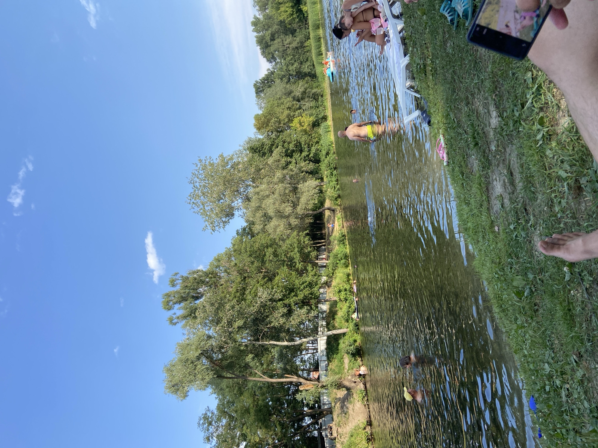 This image captures a sunny day at a riverbank in Petrivka, Ukraine. Several people are enjoying the water, with some swimming and others relaxing on the shore. In the foreground, a person's legs and feet are visible, suggesting they are sitting on the grass and holding a smartphone. The river is wide and calm, with lush green trees lining the opposite bank. In the background, there's a small blue boat with people in it, and further down the river, a few more individuals are swimming. The overall scene depicts a leisurely summer day at a popular outdoor recreational spot.