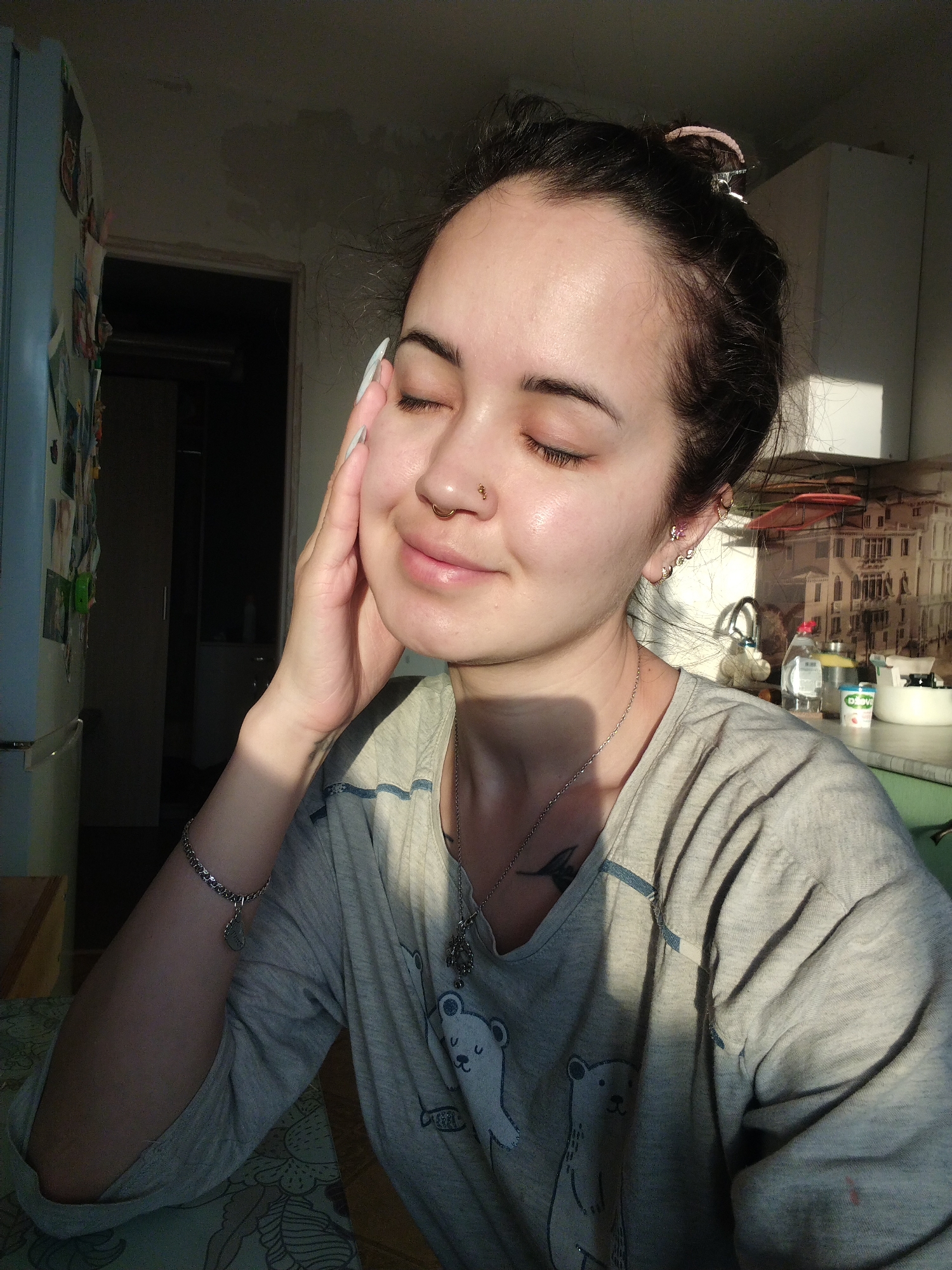 The image shows a young woman with fair skin, dark hair pulled back in a bun, and closed eyes. She is lightly smiling and appears to be enjoying the sunlight on her face. Her left hand is gently touching her cheek. She is wearing a light gray t-shirt with a cartoon polar bear design, a silver chain necklace with a pendant, and a silver bracelet on her left wrist. She has a nose ring and multiple earrings in her left ear.
The background is a domestic interior, likely a kitchen. A blue refrigerator is visible to the left, and a cluttered countertop with various items, including bottles and jars, is to the right. A partial view of what appears to be a wallpaper depicting a building is present. The overall lighting suggests a sunny day. The setting of Noviy, Russia, cannot be confirmed or denied from the image alone.