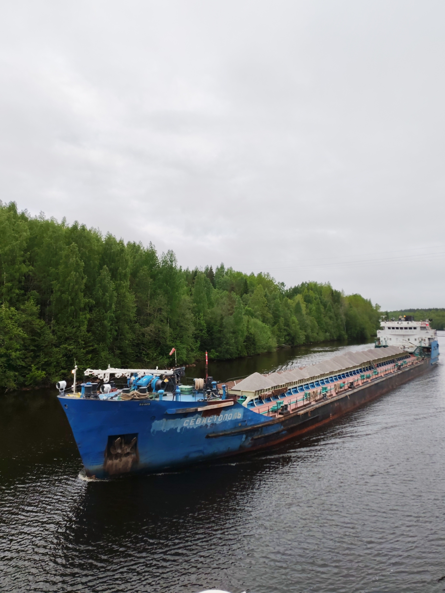 A blue barge, identified with the name "СЕВАСТОПОЛЬ" painted on its side, moves along a river or canal. The barge appears to be carrying a load of sand or similar material, covered by multiple light-colored tarpaulins. The banks of the waterway are lined with dense green trees. The sky is overcast, suggesting a cloudy day. The location is Depo, Russia, though this cannot be directly confirmed from visual elements alone.