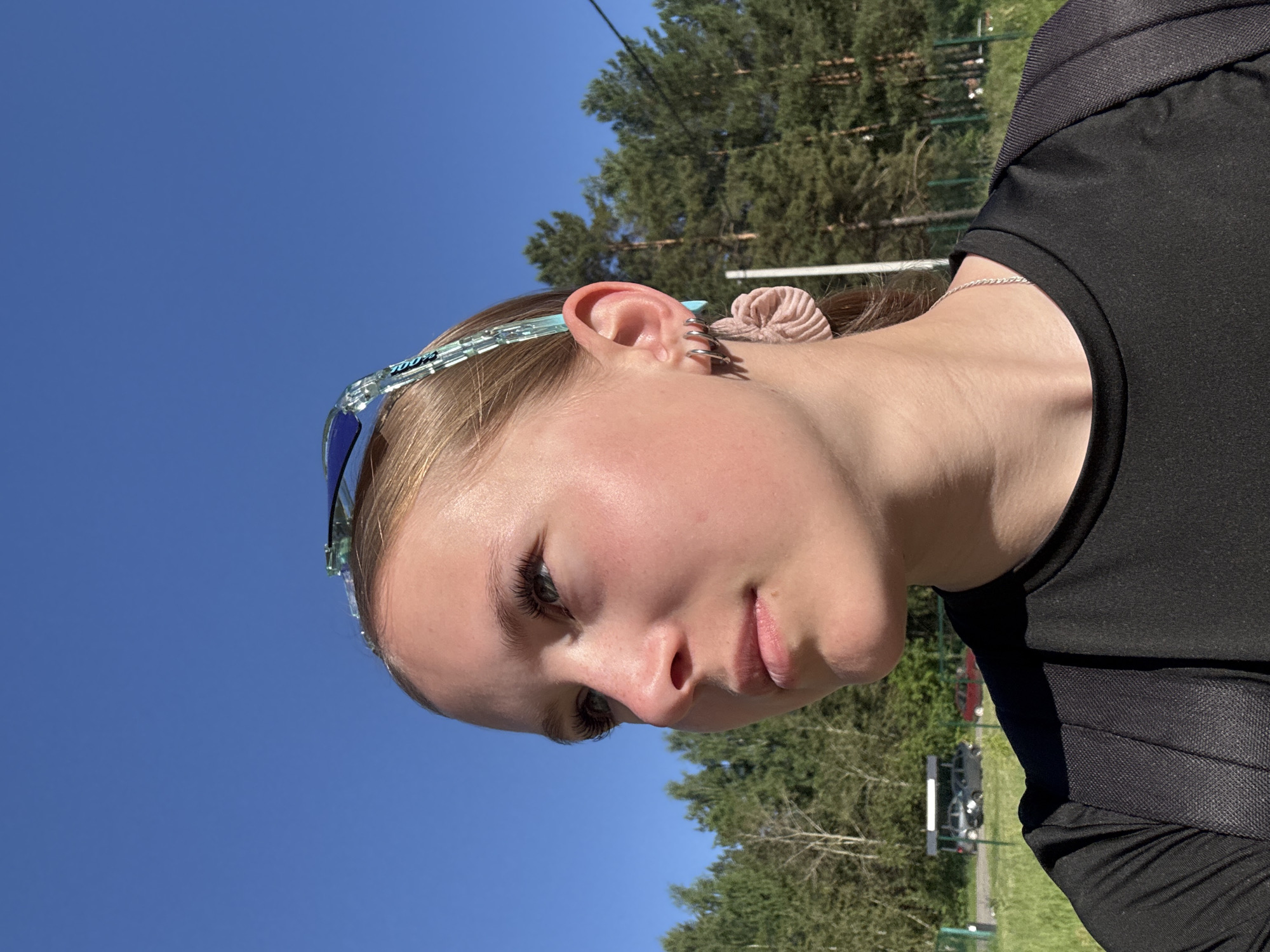 A young woman with light brown hair, wearing a black t-shirt and a backpack, poses for a portrait outdoors on a sunny day. She has on blue-tinted sunglasses pushed up on her head and wears several hoop earrings in her left ear, along with a scrunchie on her earlobe. Her gaze is directed upwards and to the left. In the background, a dense forest of pine and birch trees is visible, with a clear blue sky above. A few cars and a small building are faintly discernible in the distance, suggesting a park or recreational area, possibly near the city of Chaikovsky, Russia.