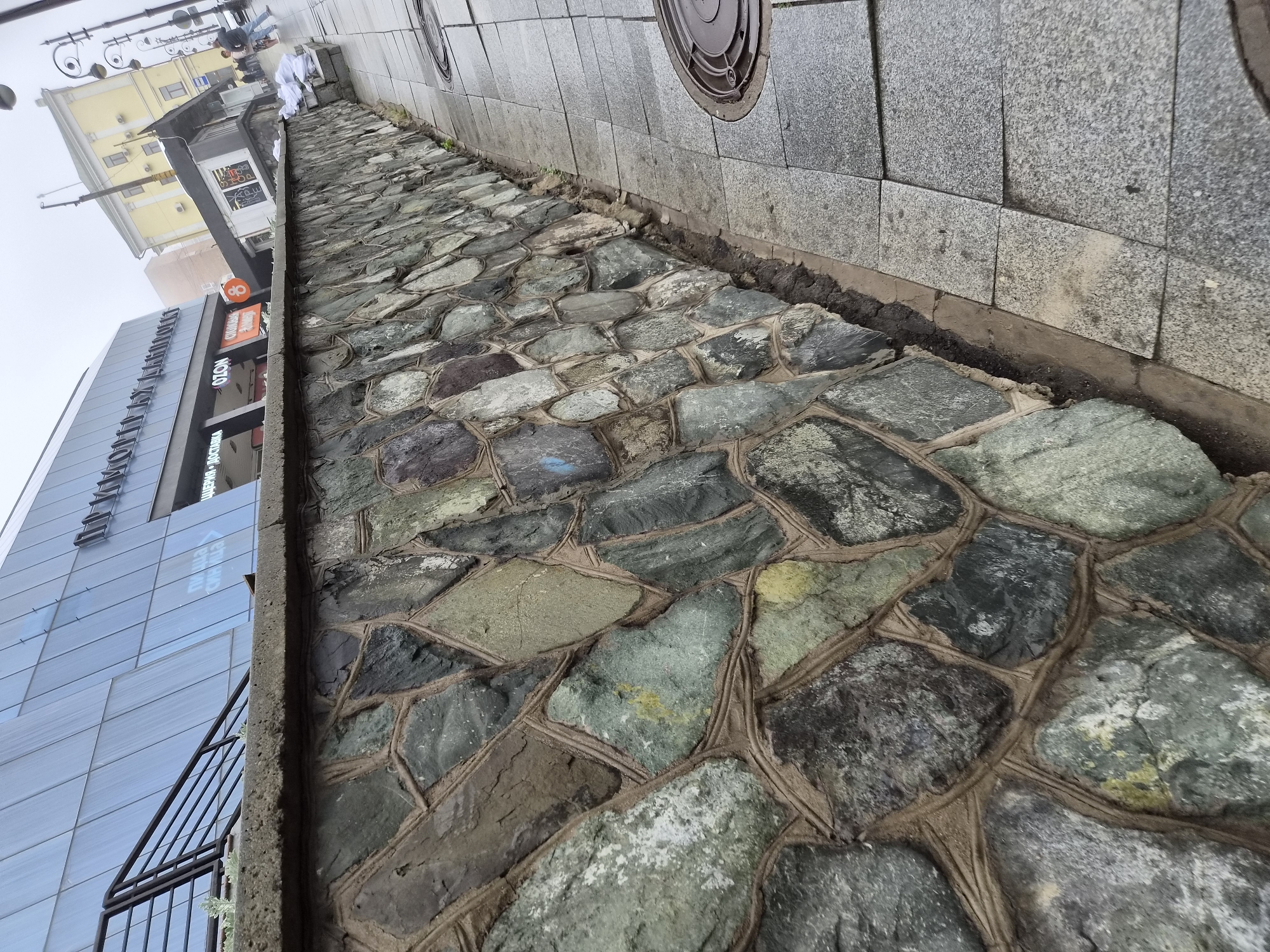 A stone retaining wall, constructed with irregularly shaped, large stones of varying grey and green hues, lined with a tan mortar, runs along the left side of the frame, angling away from the viewer. The wall separates a sidewalk from a raised area with a building. The building on the left is modern, with a blue, faceted glass facade and a black metal railing leading up to it. Text is visible on the glass facade, appearing to be in Russian, possibly reading "ПОТРЯСАЮЩУЮ КРАСОТУ". To the right of the stone wall is a grey, tiled sidewalk. In the background, a yellow building with multiple windows and a distinctive architectural style stands. Several lampposts line the sidewalk. A few people are visible walking on the sidewalk in the distance. The sky is overcast, suggesting a foggy or cloudy day. The overall scene appears to be in an urban environment, likely a public space in Vladivostok, Russia, given the Cyrillic text on the building.