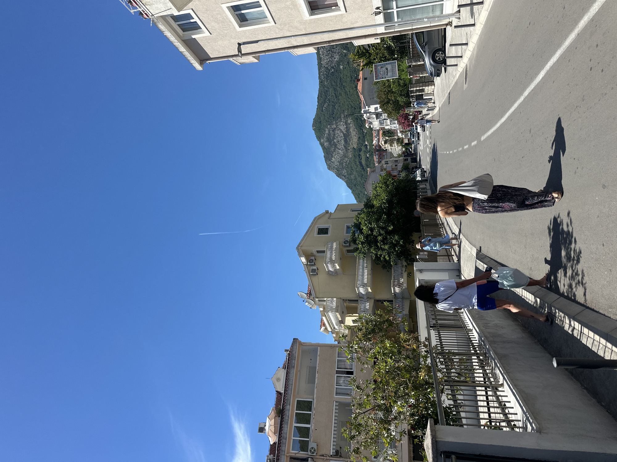 Two women walk away from the camera down a street in Budva, Montenegro. The woman in the foreground on the right wears a black bikini top and a patterned, flowing skirt. She carries a large white tote bag over her shoulder. The woman in the foreground on the left wears a white t-shirt and blue shorts. She carries a light blue shopping bag in her right hand. Both women are walking on the right side of the street, away from the viewer, on a sunny day. The street is paved and lined with buildings and trees. In the background, a large, green, mountainous hill rises above the city. The sky is bright blue with a faint airplane contrail visible. The buildings are of various heights and architectural styles, with balconies and windows. There are cars parked on the right side of the street and other pedestrians in the distance.