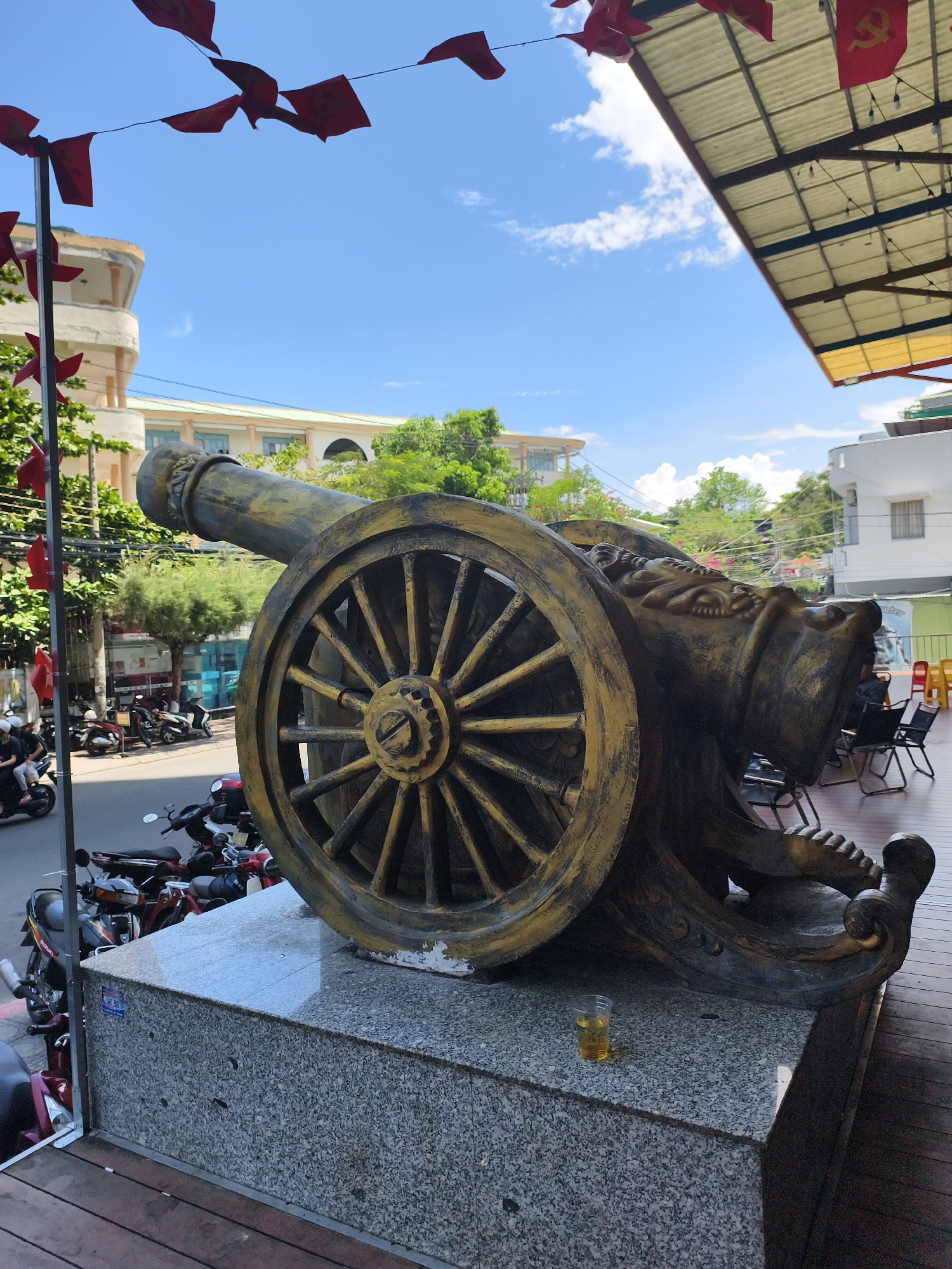 A sculpted cannon sits on a stone base in Bac Nha Trang Ward, Vietnam. The cannon's barrel, wheel, and base are bronze-colored, with visible black accents. A clear plastic cup, possibly containing beer, rests on the stone base. Red flags hang from a line above the cannon and also adorn a nearby pole. Motorbikes are parked along the street behind the cannon. A building with a curved, light-colored facade stands across the street. A covered outdoor seating area is visible to the right of the cannon. The sky is blue with scattered clouds.