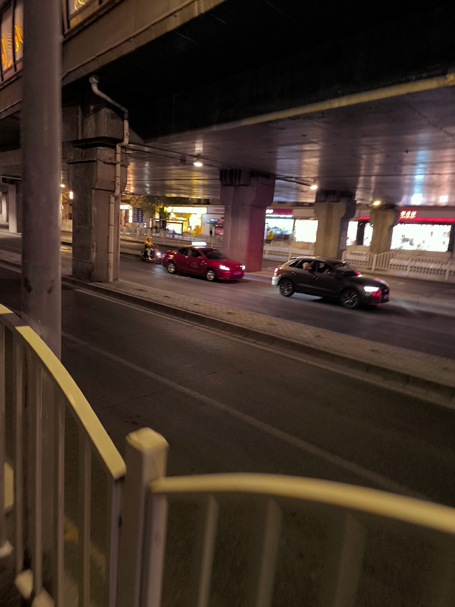 This is a night-time, medium-shot, eye-level, outdoor photograph taken from a slight angle looking down a road. The scene appears to be an urban environment, likely in Ürümqi, China, under an overpass or elevated roadway. A light-colored metal railing is in the foreground, out of focus. The road surface is asphalt with visible lane markings.
Several vehicles are on the road. Closest to the viewer, a red sedan is in motion. Further down the road, a dark-colored SUV is also moving. A scooter, possibly a delivery vehicle, with a rider wearing a yellow garment, is to the left of the red sedan. In the distance, a yellow taxi is visible.
The overpass above is constructed of concrete pillars and beams. The underside of the overpass is illuminated by overhead lights and reflections from businesses situated along the road. Some of these businesses have illuminated signage with Chinese characters. The overall lighting conditions suggest it is evening or night. The image has motion blur, indicating a moving camera or subject.