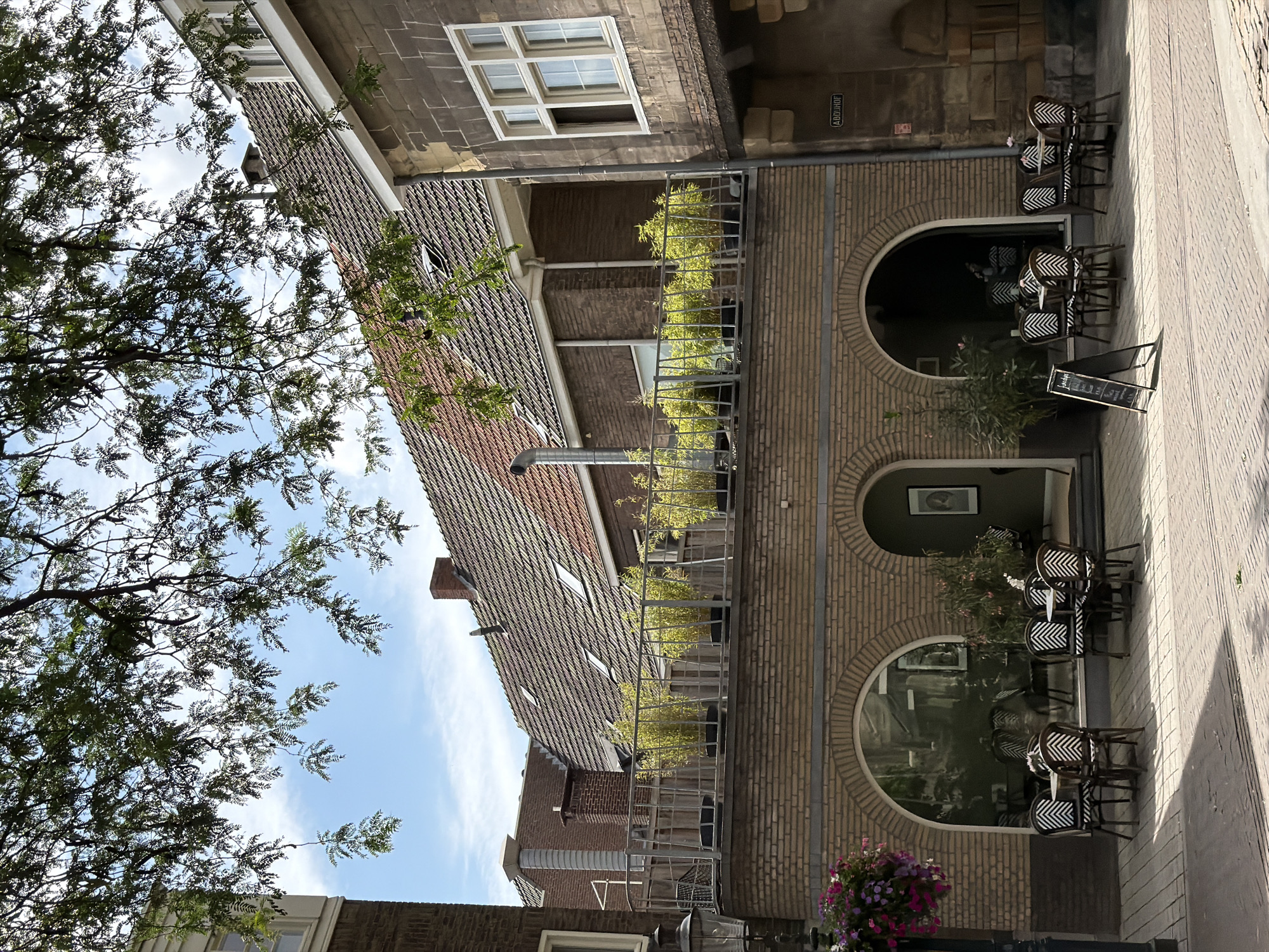 The image depicts an outdoor seating area of a cafe or restaurant in Roermond, Netherlands. The cafe is situated on a street with cobblestone paving. In the foreground, there are several tables and chairs with a black and white zigzag pattern on the upholstery. A-frame chalkboards are placed near the tables. The cafe has a brick facade with large arched windows, some of which reveal the interior. Above the cafe level, there is a balcony or terrace with metal railings, where several potted plants, likely bamboo, are displayed. In the background, there are traditional Dutch buildings with tiled roofs and dormer windows. Green foliage from a tree branches into the frame from the top, casting shadows on the scene. The sky is blue with some scattered clouds. The sign visible on the building reads "ABDHOF."