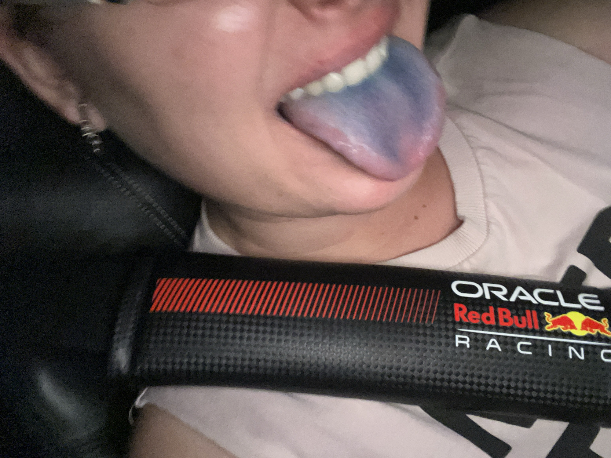The media file shows a close-up of a person's mouth with their tongue extended. The tongue is dyed a vibrant blue and purple, with hints of pink towards the tip. The person's teeth are visible and appear white. They are wearing a light-colored t-shirt and an earring in their left ear.
In the foreground, a black object with a carbon fiber texture is visible. It features red diagonal lines on the left side and text on the right, including "ORACLE," "Red Bull," and "RACING," along with the Red Bull logo. This object appears to be a branded item, possibly related to motorsports. The overall lighting suggests an indoor setting, and no specific time of day or weather conditions can be determined. The focus is on the person's mouth and the tongue's unusual coloring, creating a visually striking and somewhat unusual composition. The presence of the Red Bull branding suggests a connection to Formula 1 racing or related events.