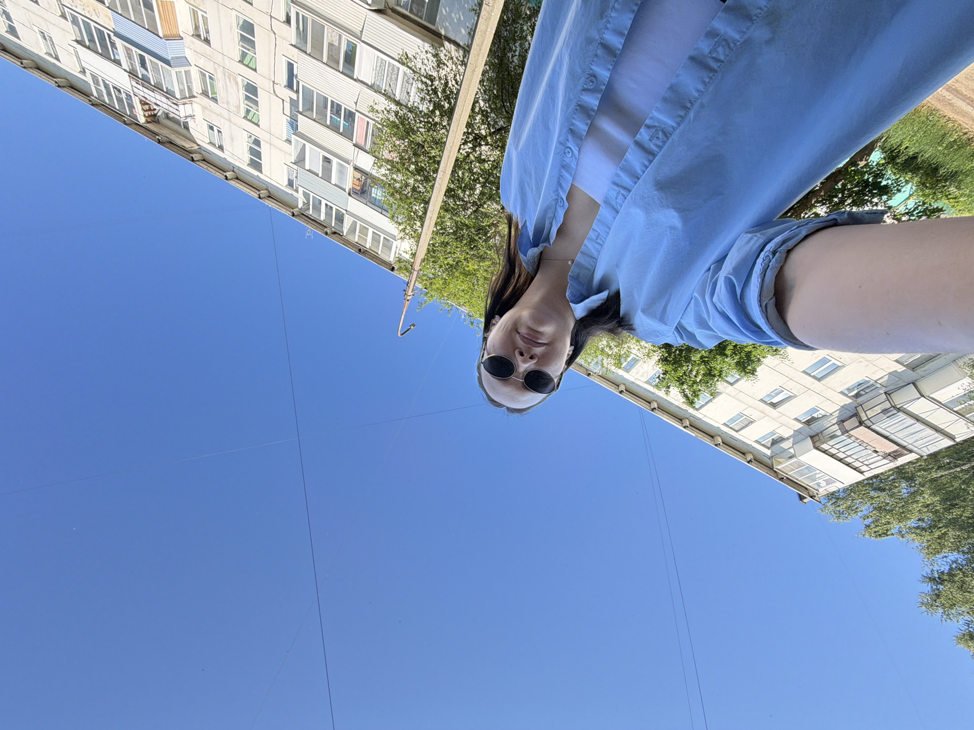 A young woman is standing outdoors and taking a selfie. She is wearing sunglasses and a light blue button-up shirt over a white tank top. Her left arm and part of her chest are visible in the foreground. In the background, there is a multi-story apartment building and trees under a clear blue sky. The image is taken from a low angle, looking up at the woman and the buildings. The location is identified as Chaikovsky, Russia.