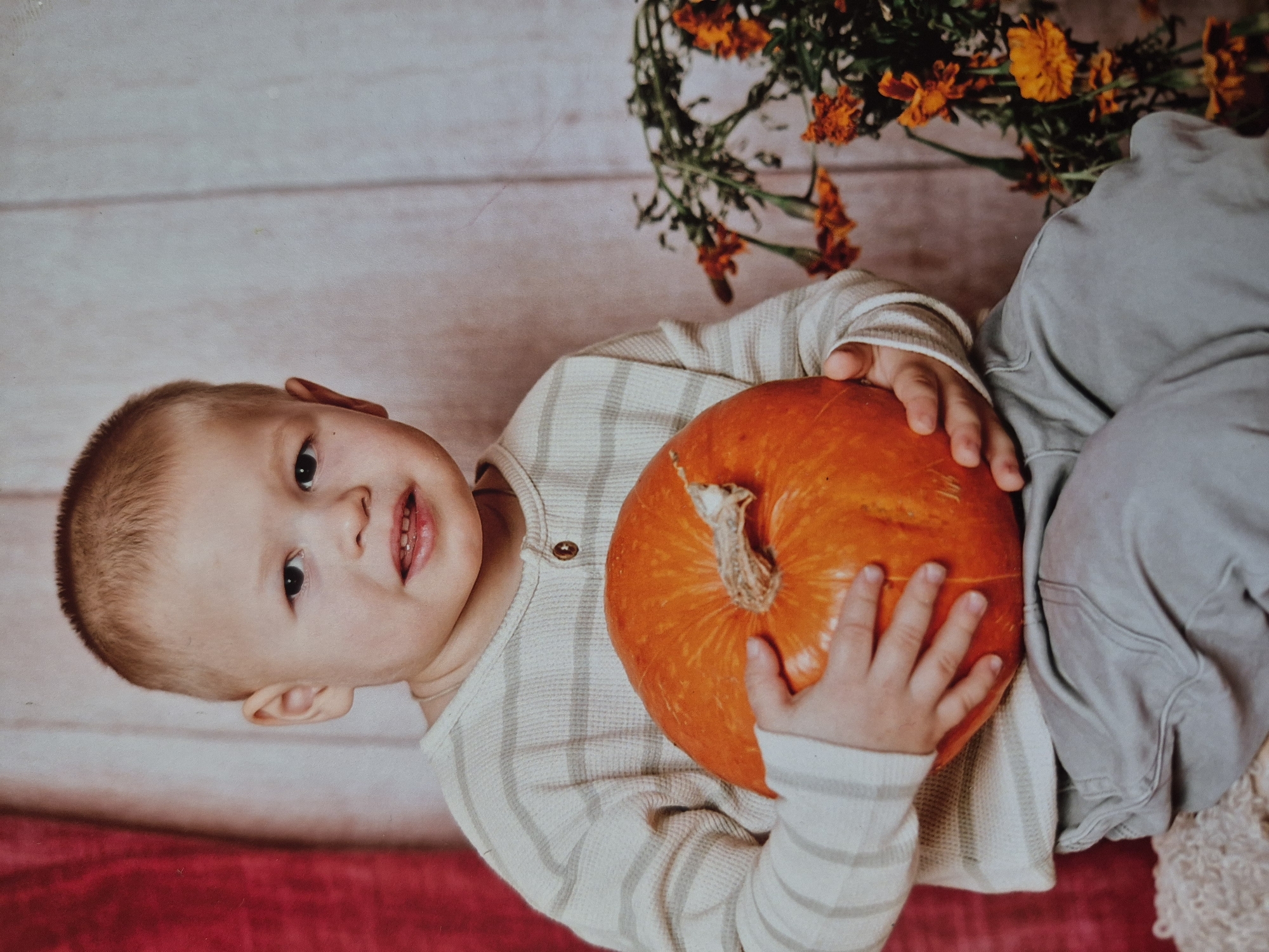 A young child is holding an orange pumpkin. The child has short, light brown hair and is wearing a long-sleeved shirt with light gray and white stripes and gray pants. The child's hands are holding the pumpkin, which is positioned in the center of the frame. Behind the child, there is a bouquet of orange flowers. The background appears to be a wall with a white and gray wood-like pattern. The photo has a warm and slightly muted color palette. The location of the city of Tobolsk, Russia is not discernible from the content of the image.