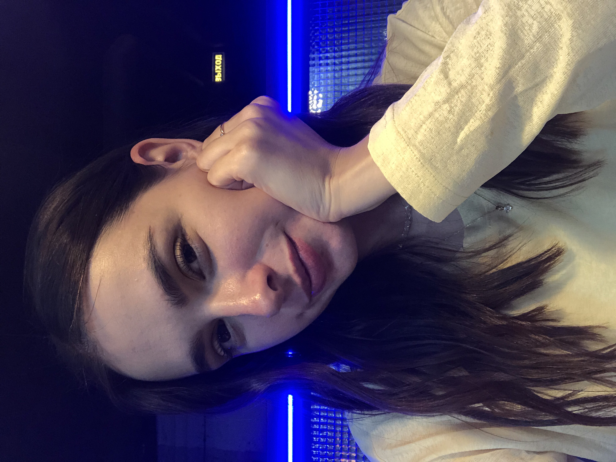 A young woman rests her cheek on her hand, her head tilted to the side. She has long, dark hair that cascades around her shoulders and down her back. Her eyes are dark and appear to be looking downwards. She is wearing a light-colored, textured top. A ring is visible on her right hand. Behind her, a blue neon light runs vertically, illuminating a textured surface. In the upper left of the image, a yellow sign with Russian text is visible. The overall lighting is dim, with the blue light being the dominant light source. The scene appears to be indoors, possibly a club or a dimly lit room. The woman's expression could be interpreted as thoughtful or relaxed.