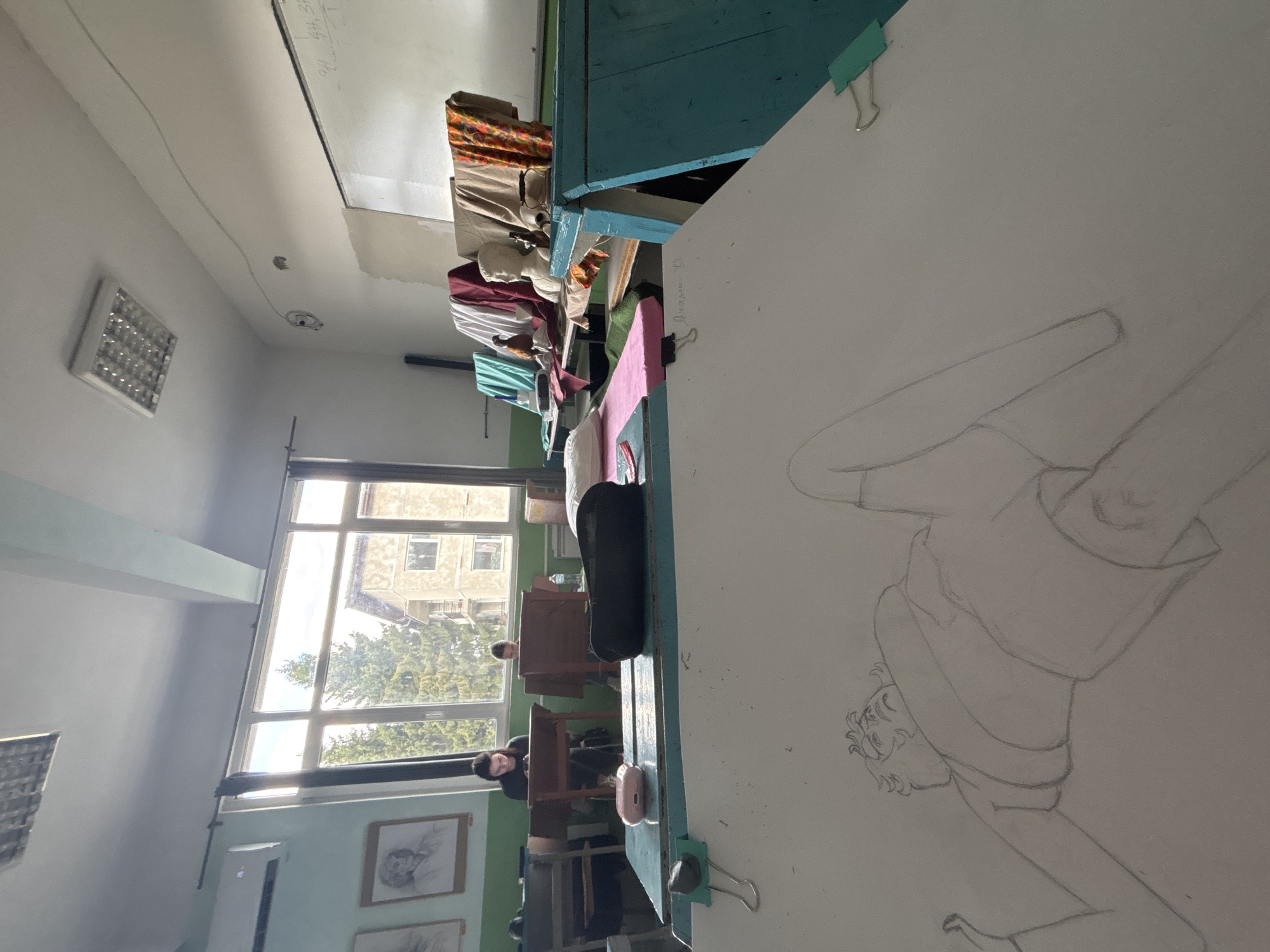 This image depicts an interior view of an art classroom or studio located in the city of Troyan, Bulgaria. The perspective is from a desk in the foreground, looking towards the windows and the back of the room.
In the immediate foreground, a white drawing board is angled towards the viewer, featuring a pencil sketch of a stylized figure, possibly female, depicted in a reclined or lying position, looking upwards. The drawing board is secured by two light-green binder clips. It rests on a light blue table, which also holds a pink earbud charging case and a dark, rectangular pencil case.
In the mid-ground, two individuals are seated at separate wooden desks. The person on the left, with dark hair and wearing a dark top, is observed looking down at a mobile device. The second person, further to the right and partially obscured, is also looking downwards. Both appear engaged in individual tasks.
Beyond the seated individuals, a large window spans the left side of the back wall, revealing an exterior view of green trees and a multi-story building under natural light. An air conditioning unit is mounted above the window. To the left of the window, two framed portrait drawings are displayed on the lower green section of the wall.
On the right side of the room, along a long counter, various still-life elements are arranged. These include several draped fabrics in colors such as teal, white, maroon, beige, and an orange patterned material. A white classical bust of a head and a dark teapot or kettle are positioned among the fabrics. A whiteboard with faint writing is visible on the far right wall.
The room features a white ceiling with a single rectangular fluorescent light fixture. The walls are painted light green on the lower half and white on the upper half. The scene is illuminated by both natural light from the window and overhead artificial lighting.