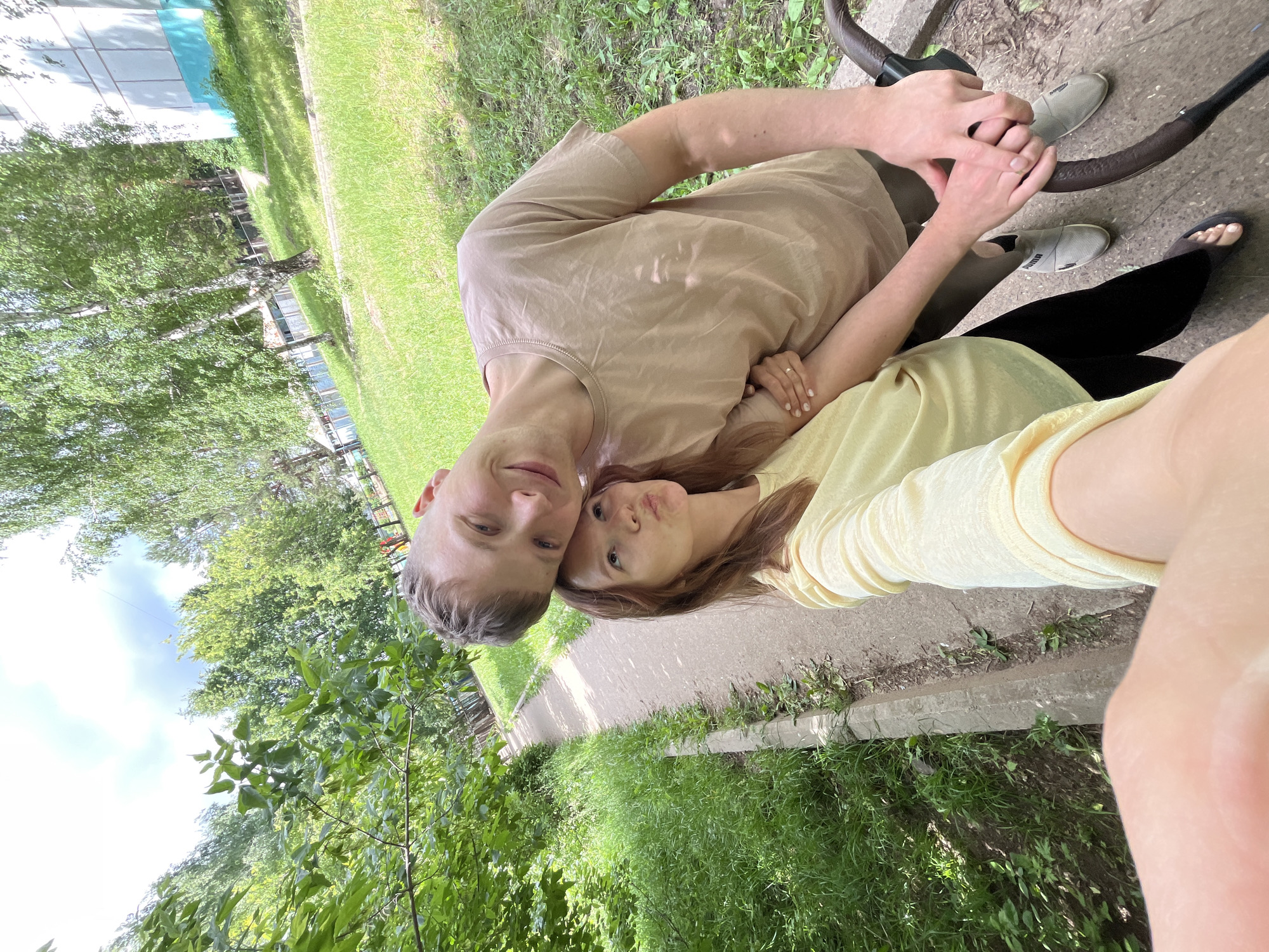 An outdoor, eye-level, wide-angle, selfie-style photograph captures a young couple standing on a paved path. The couple is positioned in the lower half of the frame, with the woman on the left and the man on the right. Both are smiling and looking towards the camera. The woman has light brown hair and is wearing a yellow top and black pants. The man has short blonde hair and is wearing a beige t-shirt and dark pants. Their hands are intertwined, and they appear to be holding onto the handle of a stroller. The background consists of a grassy area on the left and a sidewalk leading further into the distance, bordered by trees and a building. The image was taken in Chaikovsky, Russia. The overall lighting suggests it is daytime with a partly cloudy sky.