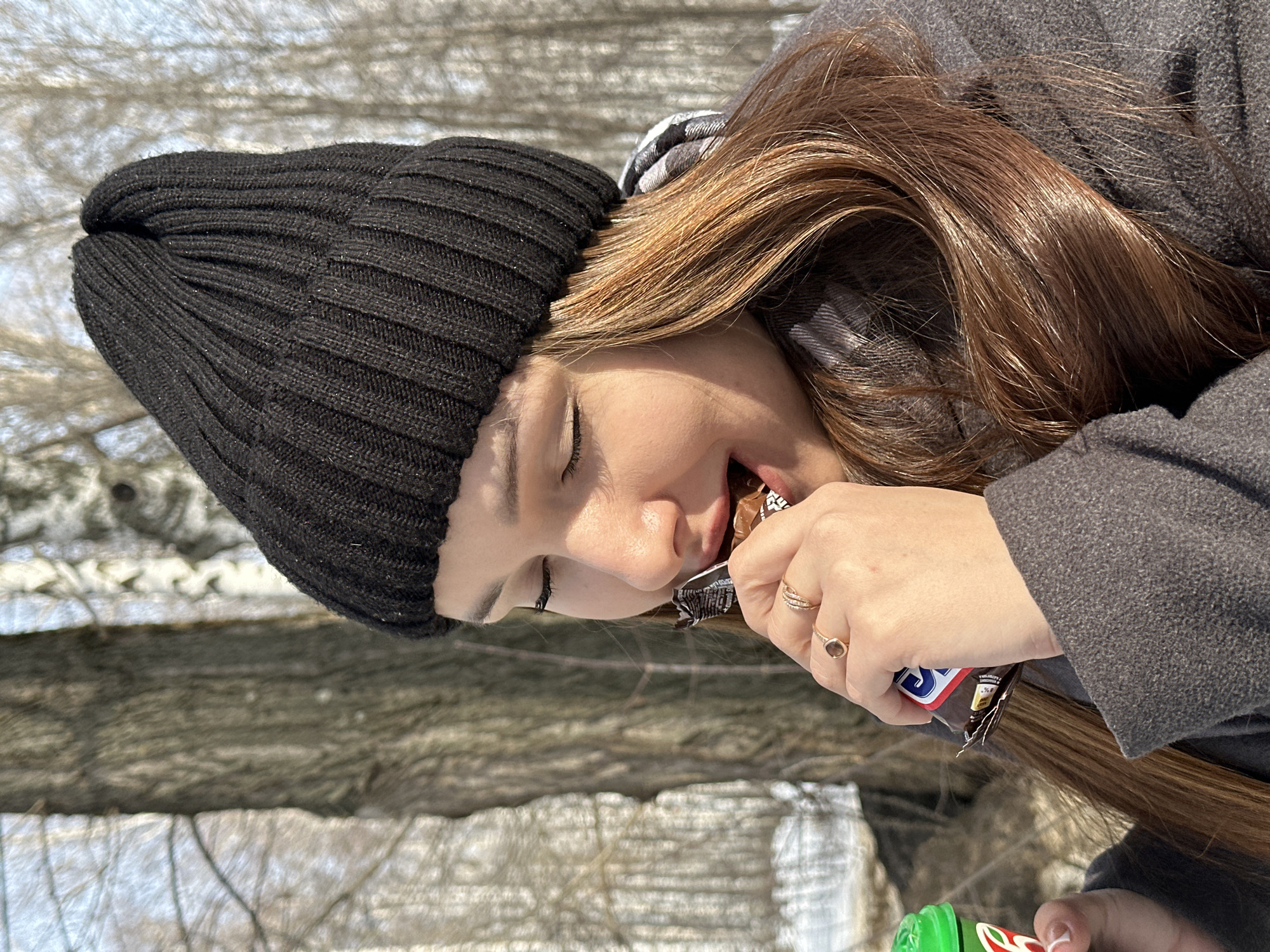 A young female individual is depicted in a close-up, chest-up outdoor portrait, engaged in consuming a chocolate bar and holding a beverage. She has closed eyes, an open mouth, and appears to be smiling. Her long, reddish-brown hair is visible below a black ribbed knit beanie. She is wearing a dark grey textured coat and a plaid scarf. Two gold-colored rings, one with a dark stone, are visible on her left ring finger. Her left hand holds a partially unwrapped "Snickers" chocolate bar, from which she is taking a bite. Her right hand holds a small green cup with a white lid, featuring a partial red and white logo, possibly for "Mountain Dew." The background depicts a sparse, leafless wooded area with multiple tree trunks, some appearing as birch, and visible snow patches on the ground, consistent with a cold season. The scene is illuminated by strong sunlight. This outdoor setting is located in Chaikovsky, Russia; however, a specific street or park within the city is not visually identifiable.