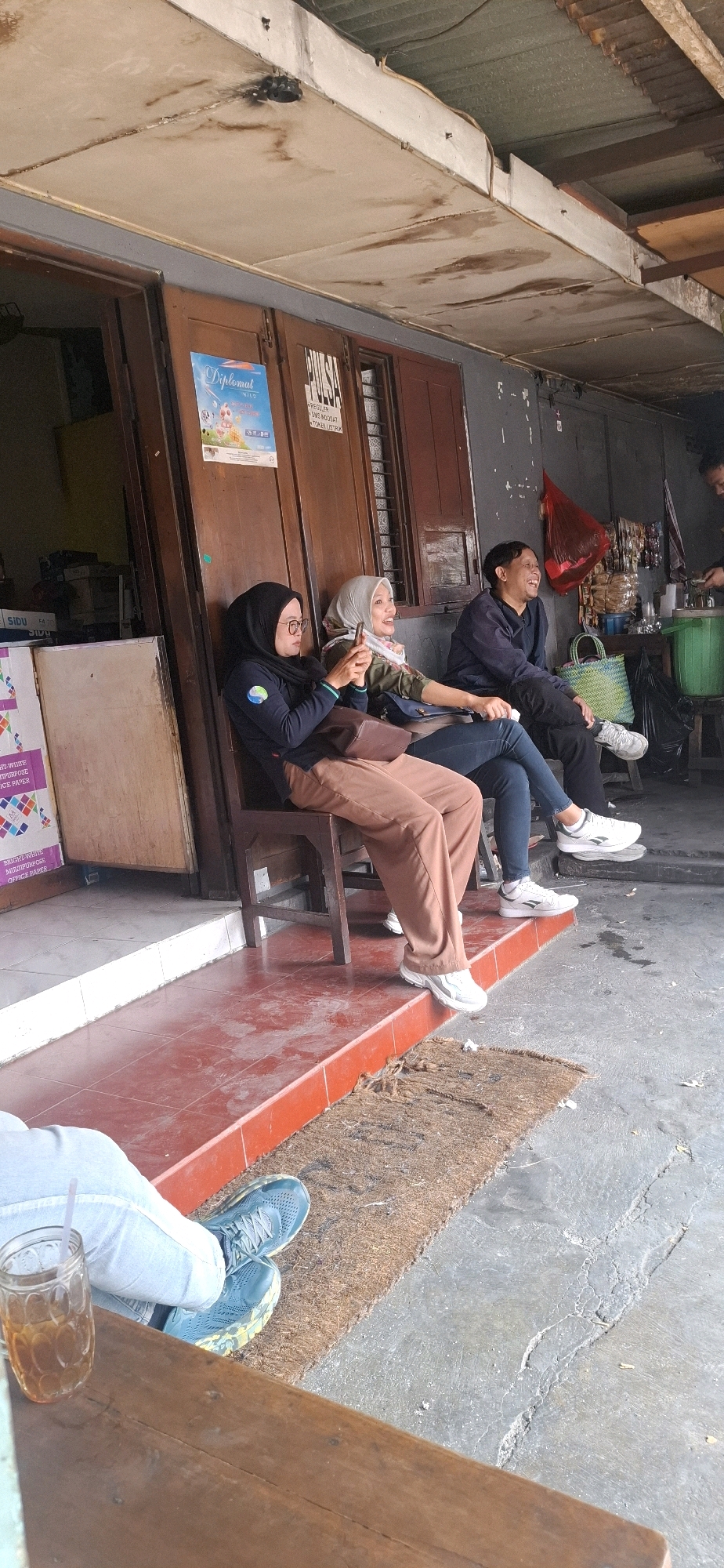 The image depicts an outdoor seating area outside a shop in Surakarta, Indonesia. Three individuals are seated on chairs, with a fourth person partially visible in the background. The first person, wearing a black hijab and glasses, is seated on the left, holding a smartphone. To their right, a person in a light-colored hijab is also looking at their phone and smiling. The third person, a man in a dark blue long-sleeved shirt, is laughing and looking to the right. In the background, a person is tending to some items on a counter. The shop appears to be a small business, possibly a convenience store, with signs visible on the doors, including one that reads "Diplomat NILS" and another with "PULSA". The environment is somewhat worn, with a corrugated metal roof, peeling paint on the walls, and a tiled floor. A glass of iced tea with a straw is visible in the foreground on a wooden surface. The overall impression is a casual gathering outside a local shop.