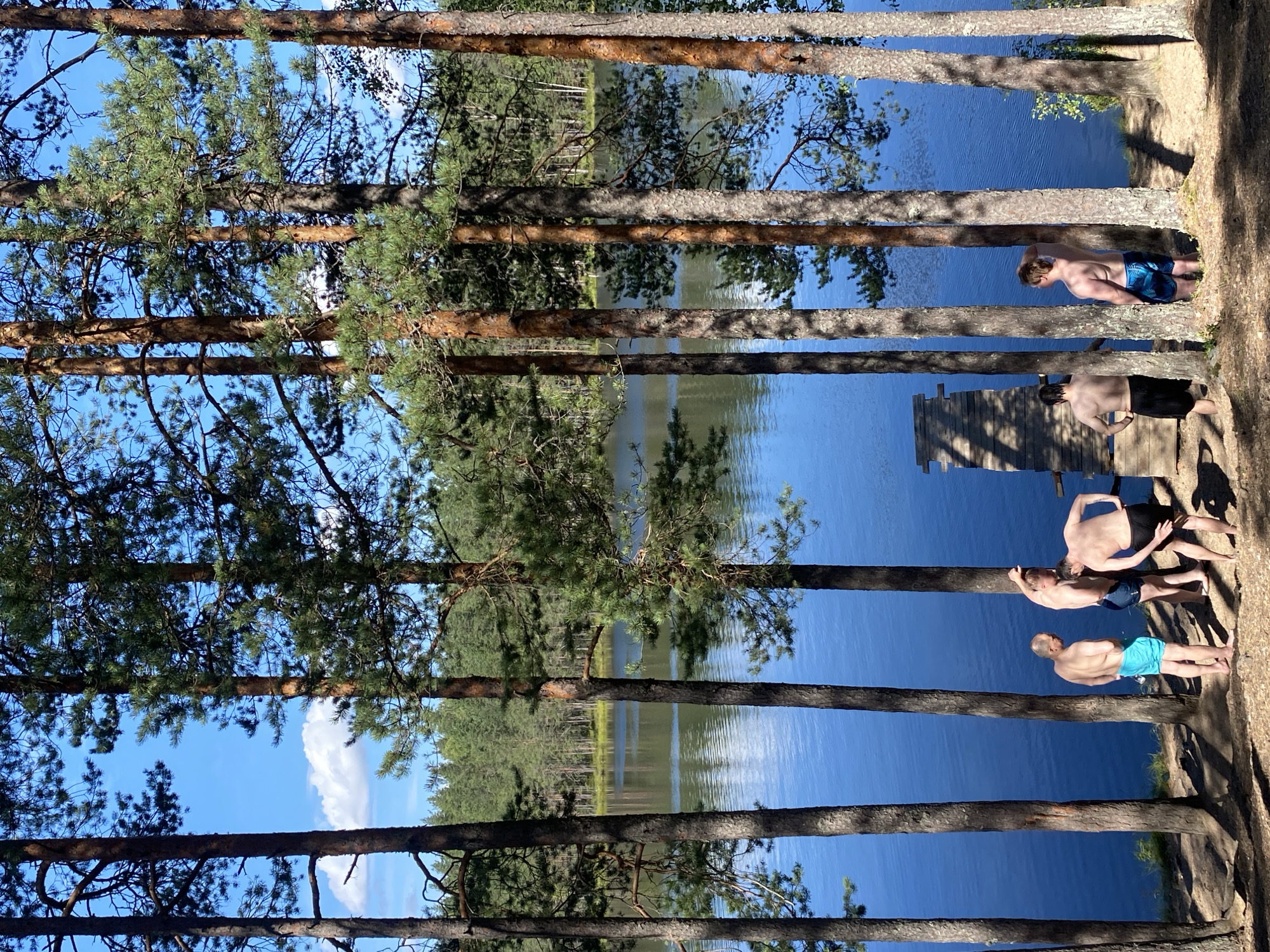 In Leipyasuo, Russia, a group of men are gathered near a lake. The scene is set amongst tall pine trees, with the deep blue water of the lake visible beyond. The men, all shirtless and wearing swim trunks, are positioned on a dirt embankment. One man in blue swim trunks stands to the left, facing away from the camera. To his right, two men are in conversation, one with blonde hair and wearing dark blue trunks, the other with dark hair and black trunks. Further to the right, another man in blue and black patterned swim trunks stands with his back to the camera, looking out at the water. A small wooden pier is visible on the left side of the frame, extending into the lake. The sky is clear and blue with a few scattered white clouds, and sunlight casts shadows through the trees onto the ground.
