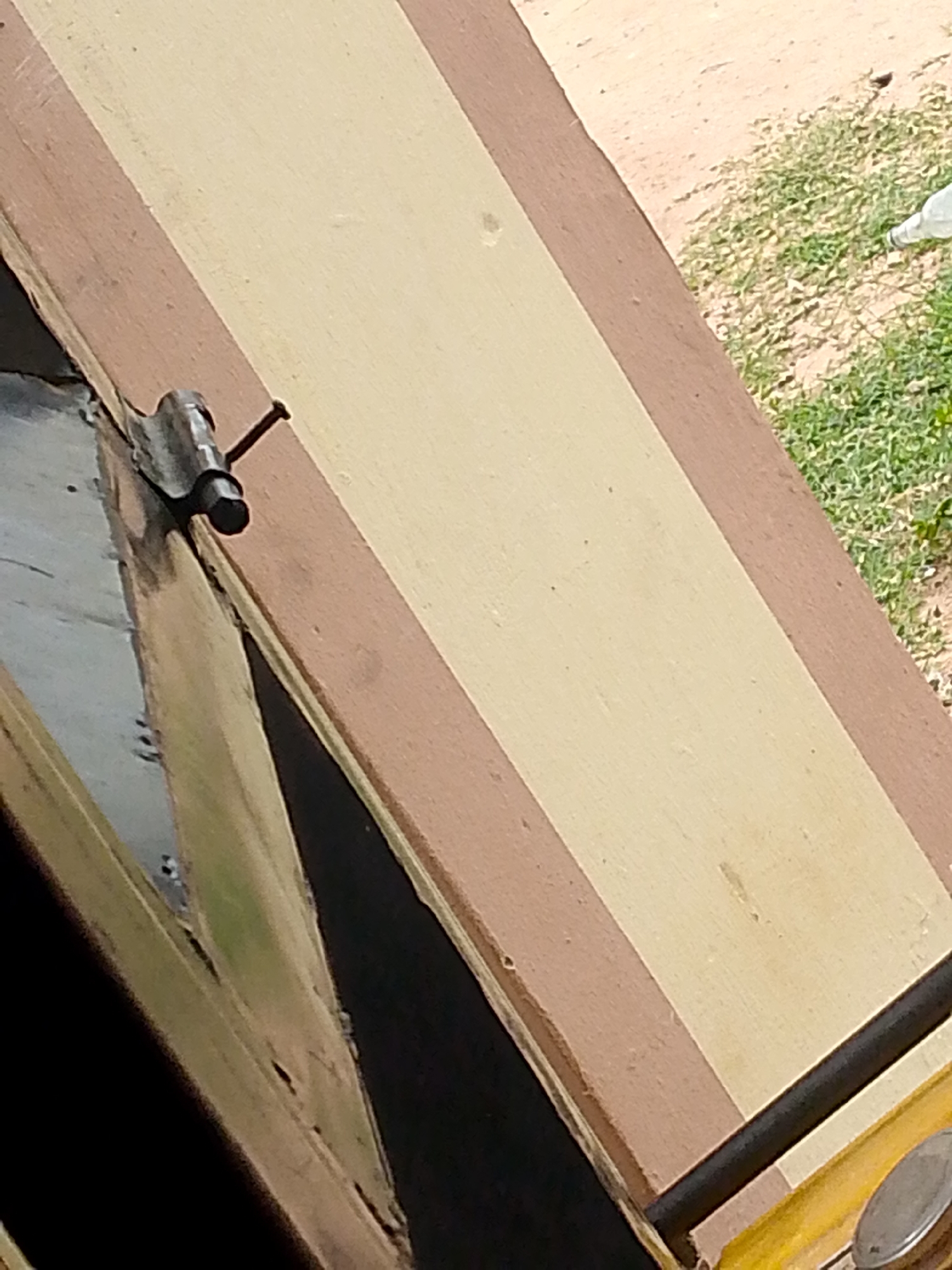 In Jauro Yinu, Nigeria, a close-up shot reveals a metal bolt and nail on a wooden surface, divided into bands of brown and cream. The bolt is latched, its handle protruding outwards. A nail, bent at an angle, is hammered into the wood near the bolt. The background shows a patch of dry dirt and sparse green grass, with a white object, possibly a bottle, lying on the ground. The lower portion of the image displays a dark, reflective surface, likely wood or metal, with some visible wear and tear.