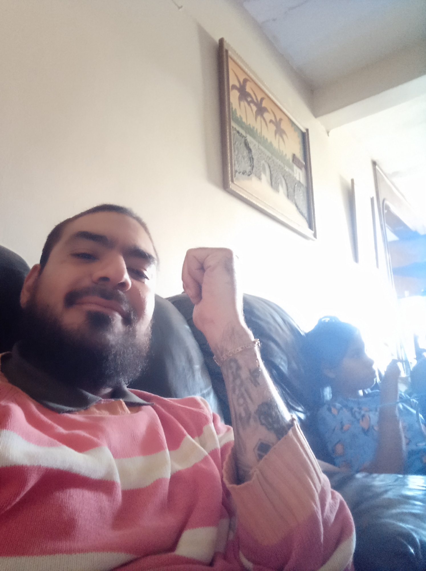 An indoor scene, likely a living room in Guarenas, Venezuela, shows a man and a child relaxing on a dark-colored sofa. In the foreground, a man with a dark beard and mustache, wearing a pink and white striped long-sleeved shirt with a dark collar, looks towards the camera with a slight, friendly smile. His left forearm, which is resting on his knee, displays several tattoos and a gold-colored bracelet, with his hand slightly clenched.
Behind and to the right of the man, a child is partially visible and out of focus. She has dark hair, seemingly tied back, and is wearing a blue patterned top. She appears to be looking towards the right side of the frame, perhaps at a television or a window.
The background features a plain, light-colored wall with framed artwork. A prominent painting depicts a stylized landscape with several palm trees and a grey, textured structure resembling a bridge or wall. This artwork is housed in a golden frame. Another framed item is partially visible further to the right. The room is brightly lit, particularly on the right side, suggesting strong natural light from a window, indicating it is daytime. The overall mood is relaxed and casual. No visible text is present in the image.