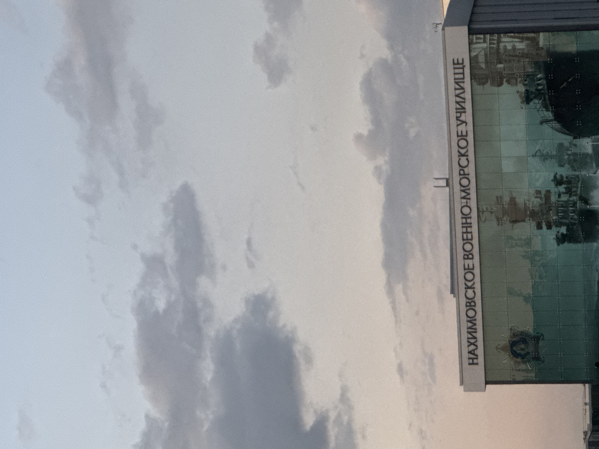 The image shows the Nakhimov Naval Academy building in Mariupol, Ukraine. The building has large glass windows that reflect the sky and images of naval ships. Text above the windows reads "НАХИМОВСКОЕ ВОЕННО-МОРСКОЕ УЧИЛИЩЕ" (Nakhimov Naval School). A crest is visible on the left side of the building's facade. The sky is cloudy, with a mix of light and dark gray clouds against a pale blue and pinkish hue, suggesting it is either early morning or late evening. The overall scene depicts a building that appears to be intact against a backdrop of a cloudy sky.