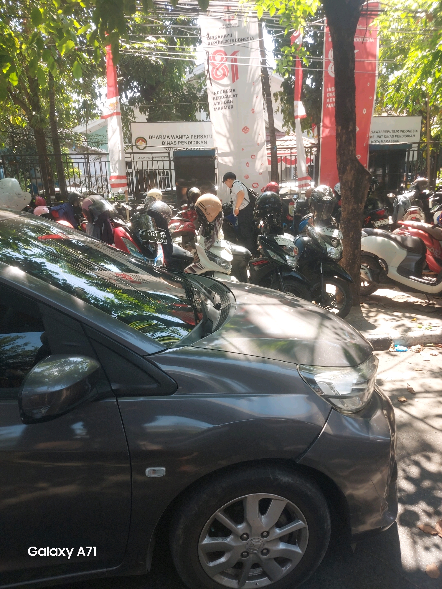 A dark gray car is parked in front of a building entrance in Makassar, Indonesia. The car is in the foreground, with its front bumper and wheel visible. Behind the car, several motorcycles are parked, some with riders. Large banners are hanging on the building, some with Indonesian text and national symbols. The area appears to be an outdoor parking lot with trees providing shade. The overall scene suggests a busy street or public space in a city.
