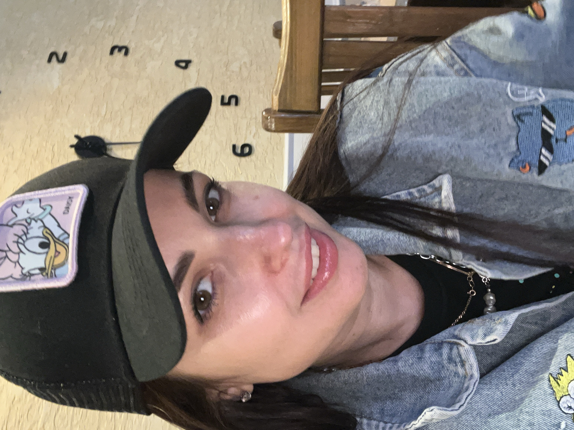 A woman with brown hair is shown from the chest up, wearing a black baseball cap with a Daisy Duck patch, a black t-shirt, a silver chain necklace, a light blue denim jacket, and diamond stud earrings. She is smiling. Behind her is a wall with a stylized clock design featuring the numbers 2 through 6, and the top of a wooden chair is visible. The location is Unknown, Unknown.