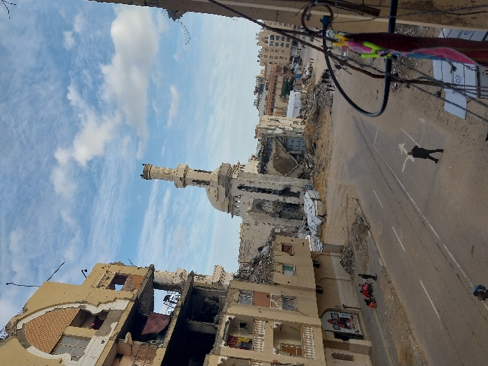 The image portrays a scene of extensive urban destruction in Khan Yunis, Palestinian Territory, under a partly cloudy blue sky. A multi-story building on the left exhibits severe damage, with large sections of its facade and internal structures missing, exposing multiple floors. Below it, on the roadside curb, a group of approximately three to four individuals is seated.
Central to the scene, further down a paved street, stands a severely damaged mosque with its minaret still largely upright but showing structural degradation and missing sections; its dome and adjacent structures are partially collapsed amidst significant piles of rubble. The ground around the mosque and other surrounding areas is covered in debris from collapsed buildings.
A single male figure in dark attire walks in the middle of the street, facing away from the camera, near a painted white directional arrow. Further along the street on the right, a group of approximately five to seven individuals can be seen walking amidst the rubble. The foreground right includes electrical wires and a clothesline with fabric items, indicating the photograph was taken from an elevated position. The background shows additional damaged buildings and debris, with what appear to be temporary structures.