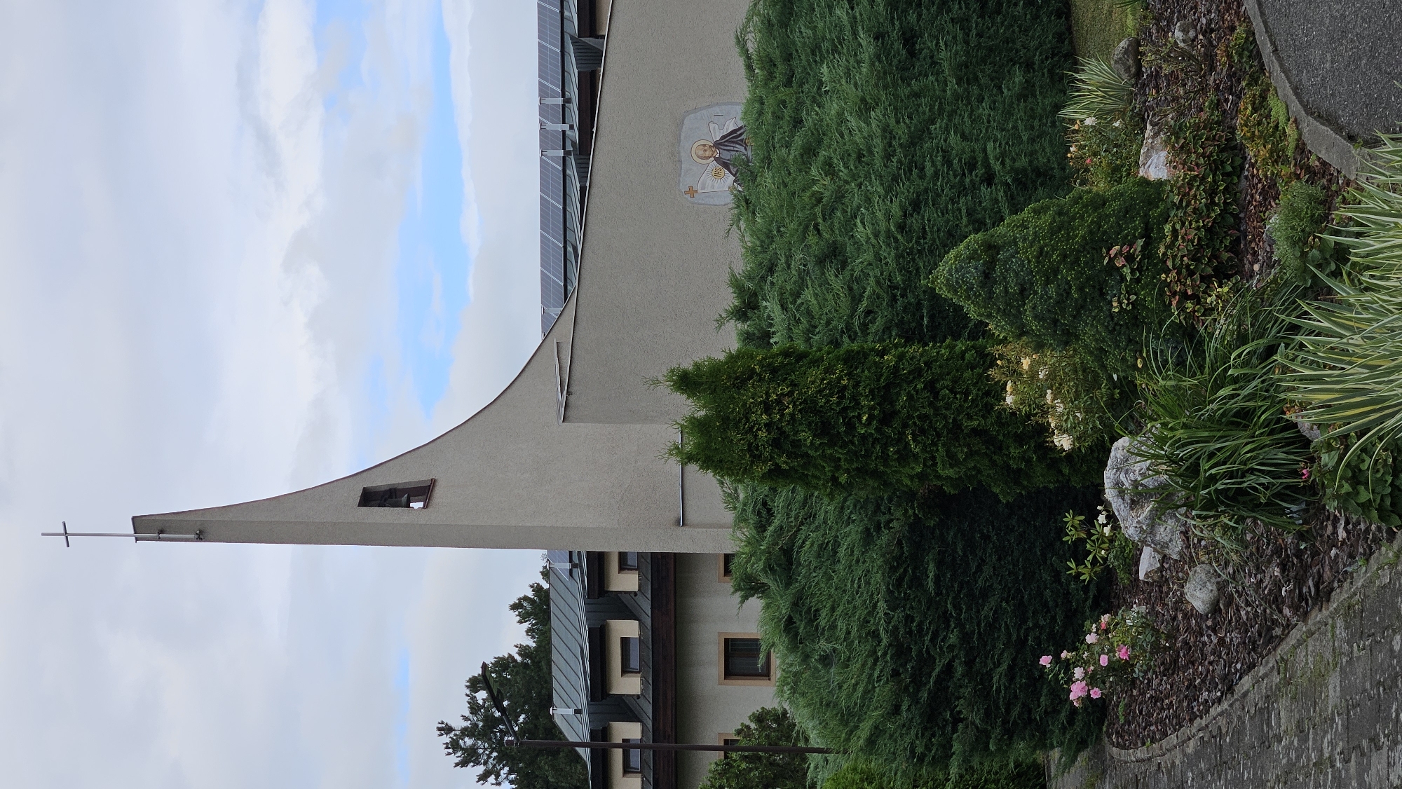 The image shows a modern church building with a tall, slender bell tower topped with a cross. Solar panels are installed on the roof of a connected building. A mural depicting a religious figure, possibly Saint John, is visible on the exterior wall. The church is surrounded by lush green bushes, trees, and a well-maintained garden with flowers and rocks. A paved walkway is present in the foreground. The sky is cloudy. The scene is likely located in Prešov, Slovakia, given the architectural style and religious iconography common in the region.