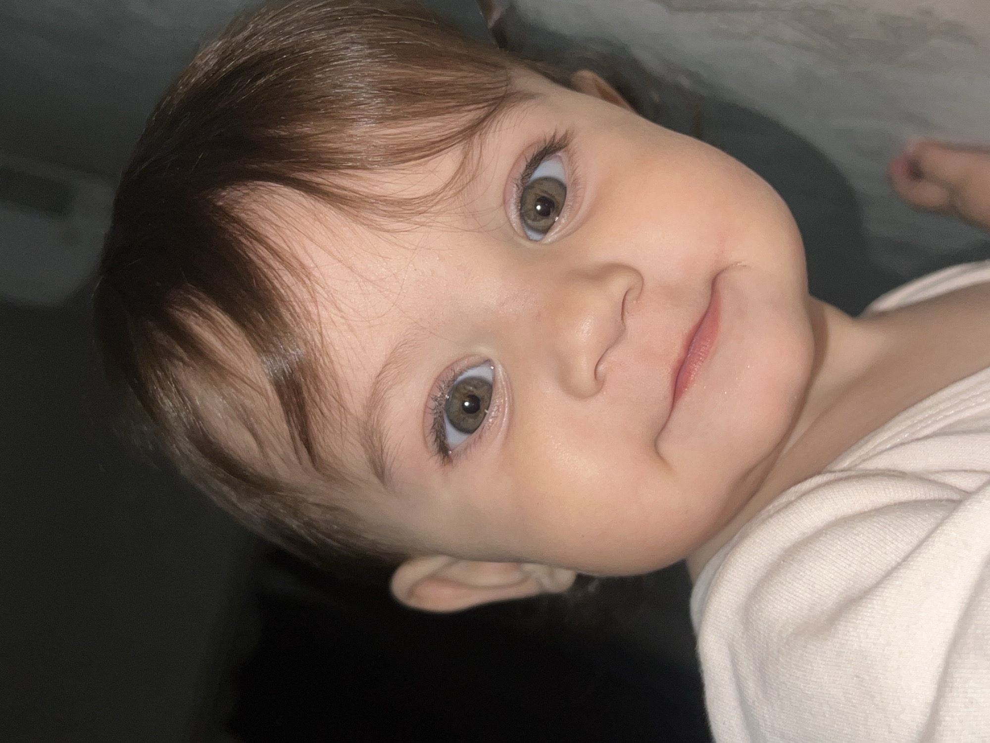 A close-up, angled portrait shows a baby with brown hair and hazel eyes. The baby's skin is fair. The child's lips are slightly parted with a hint of pink. The baby is wearing a white shirt. The background is blurry and indistinct, with darker areas suggesting a shadowed room. There is no evidence to support that this image was taken in the city of Elena, Bulgaria.