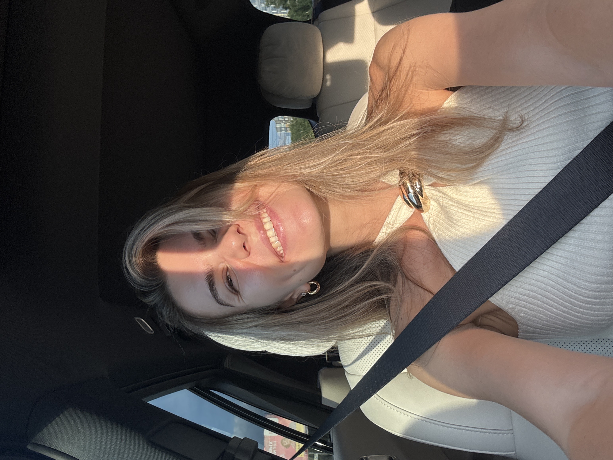 A woman with blonde hair smiles and looks at the camera from the passenger seat of a car. She is wearing a white, ribbed top with a gold clasp around the neckline and a seatbelt. Sunlight streams in from the right, casting a shadow across her face. The interior of the car is visible, including white leather seats and the headrests. Outside the car window, buildings and trees can be seen.