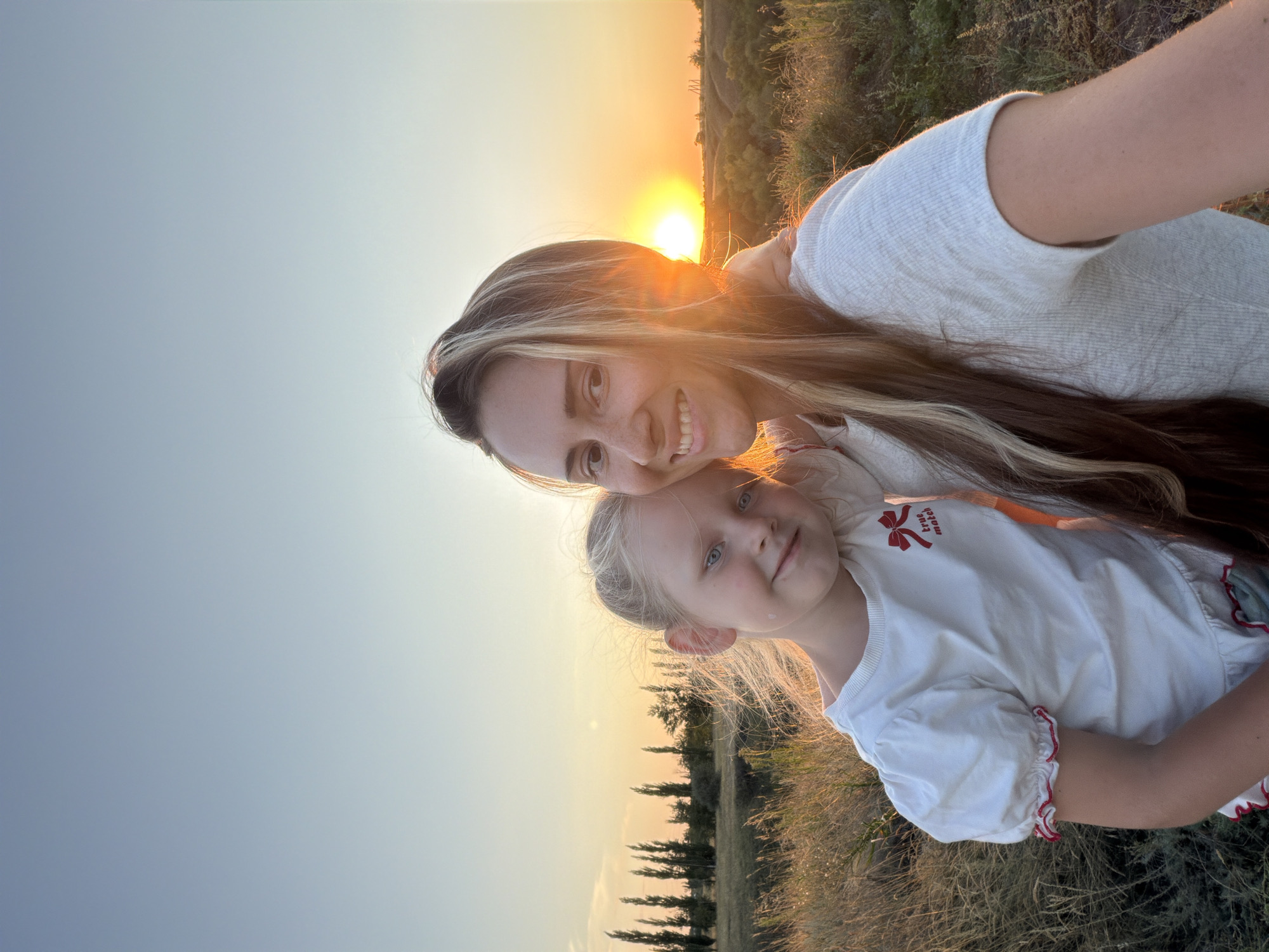 In a selfie taken at sunset in Kamyshin, Russia, a woman and a young girl are positioned in the foreground. The woman, on the right, has long blonde and brown hair and is smiling. A young girl with blonde hair and blue eyes is leaning against the woman on the left, also smiling. They are in an outdoor setting with dry grass and trees in the background. The sun is setting on the horizon, casting a warm glow over the scene. The woman is wearing a white ribbed shirt, and the girl is wearing a white puff-sleeved shirt with a red bow and the words "true match" printed on it.
