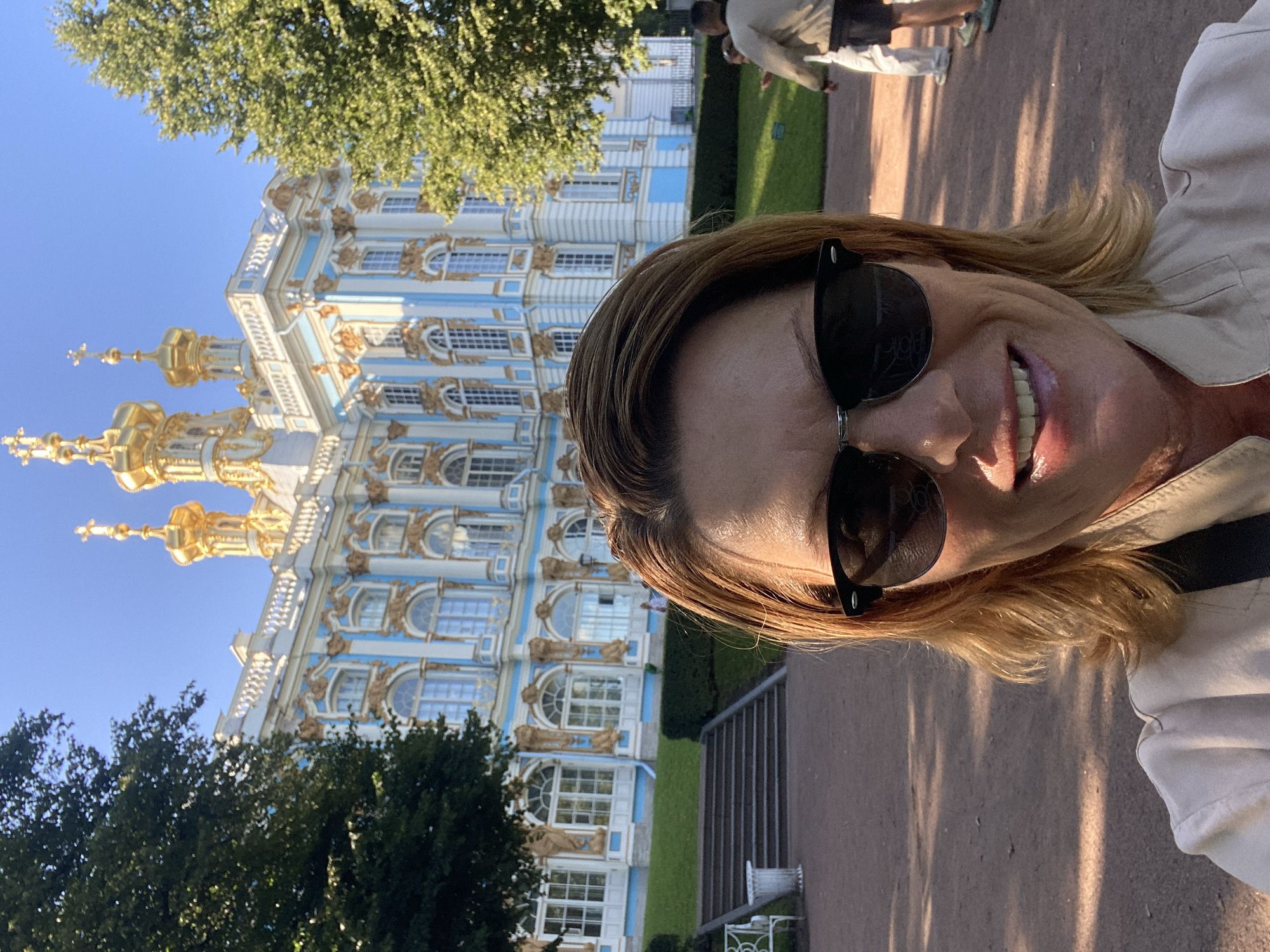 A woman with blonde hair and sunglasses smiles for a selfie in front of the Catherine Palace in Pushkin, Russia. The palace, a striking example of Baroque architecture, features blue and white facades adorned with gold accents and ornate detailing. Golden domes and spires rise majestically against a clear blue sky. Lush green trees flank the palace, casting dappled shadows on the manicured lawn and gravel pathway in the foreground. The woman is wearing a light-colored jacket. In the background, two individuals are walking on the pathway. The scene conveys a sense of sunny day tourism at a historic landmark.