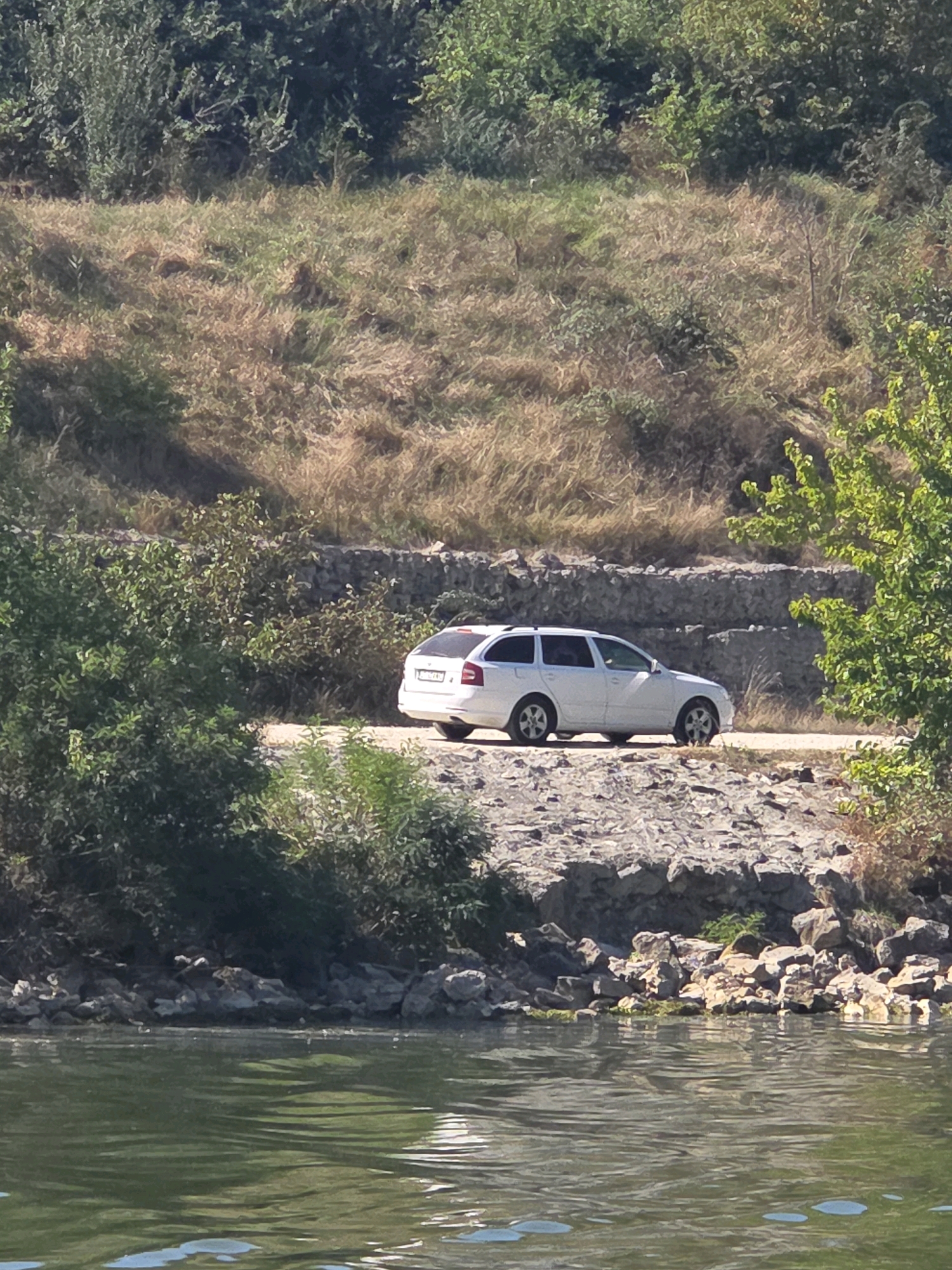 An eye-level, medium shot captures a white station wagon parked on a gravel path alongside a body of water. The car is oriented to the right, facing away from the camera. The path is bordered by a rocky embankment, and further beyond, a vegetated hillside rises. The water in the foreground exhibits reflections and ripples. The overall scene depicts a naturalistic setting, possibly near a river or lake, in Medgidia, Romania.