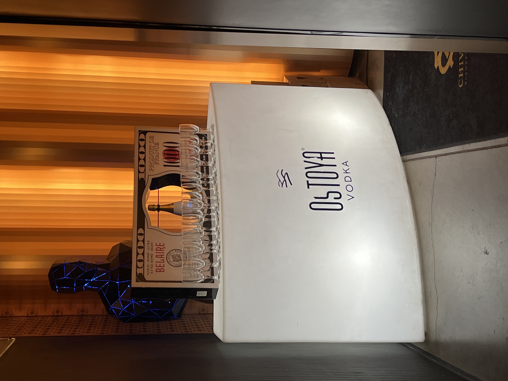 The image is a close-up shot of a promotional display for Ostoya Vodka, situated indoors. The display is a white, curved structure with the brand name prominently displayed in black lettering. To the left of the display, there is a backlit bottle of Belaire sparkling wine, set in a holder that resembles an abstract human figure with blue LED lights outlining its form. Numerous champagne flutes are lined up along the top edge of the wine bottle display. The background consists of horizontal, orange-toned blinds. A black mat with "Chivas" printed on it is visible behind the vodka display. The scene gives the impression of a bar or promotional event setup, but the precise location in Burgas, Bulgaria, cannot be determined from the image alone.