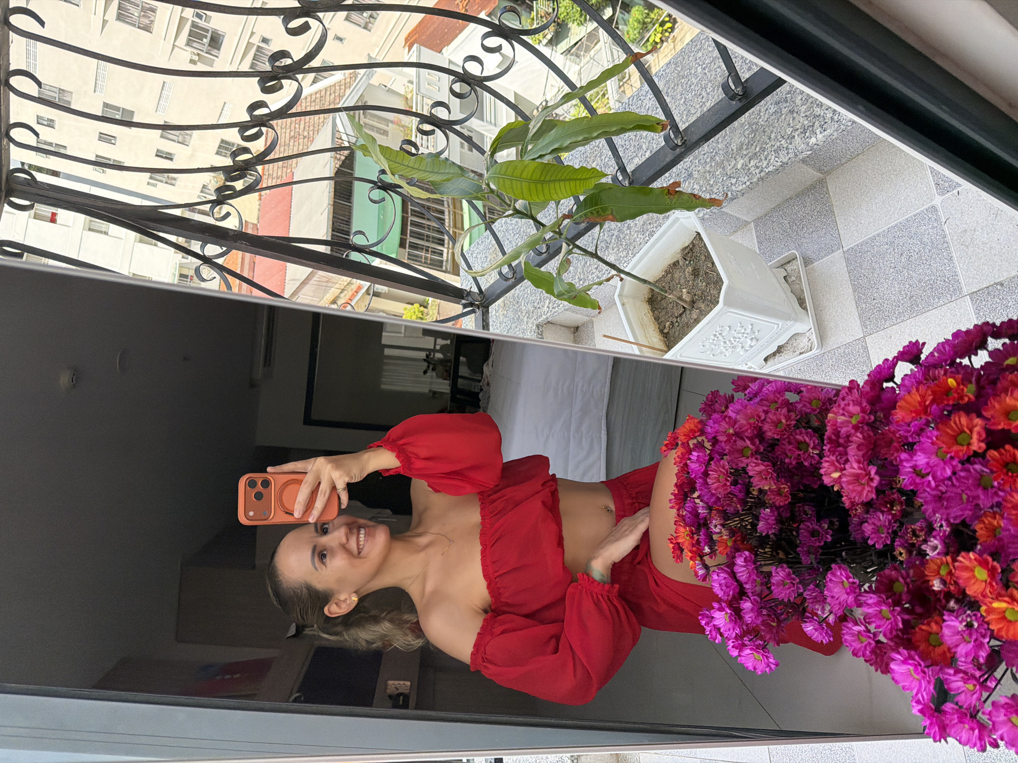 A woman is taking a selfie in a mirror on a balcony in Nha Trang Ward, Vietnam. She is wearing a red off-the-shoulder top and red shorts. In the foreground, there is a bouquet of pink and orange flowers. In the background, a plant in a white pot sits on the balcony railing, with buildings and a street visible beyond.