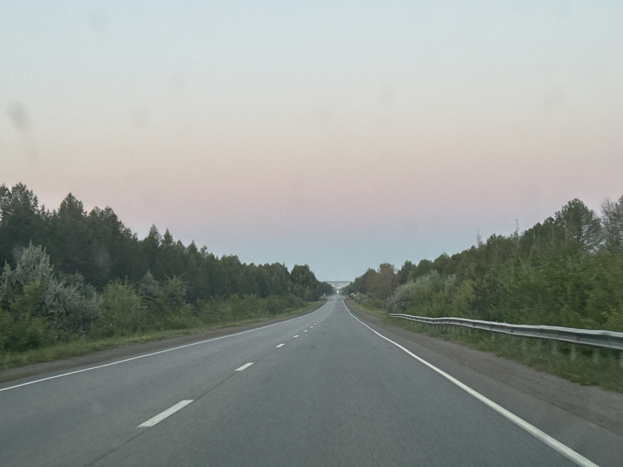 The image depicts a two-lane asphalt road extending into the distance, flanked by dense green foliage and trees on both sides. The sky above is a gradient of pale blue, transitioning to soft pink and light gray, suggesting either sunrise or sunset. White lane markings are visible on the road surface. A metal guardrail runs along the right side of the road. The scene appears to be in a rural or semi-rural area, possibly on the outskirts of Arsinskii, Russia, given the type of road and natural surroundings. There are no people or vehicles immediately visible in the frame.