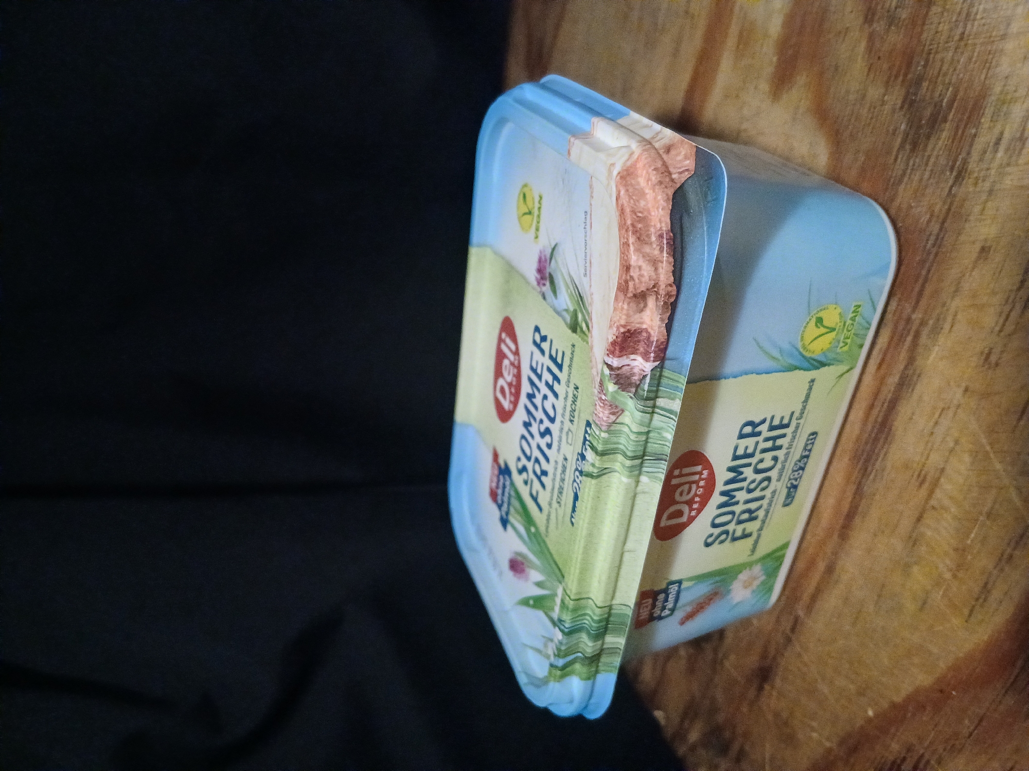 The image displays a close-up, eye-level shot of a container of "Deli REFORM SOMMER FRISCHE" spread, placed on a wooden surface against a dark background. The container is light blue with a label that features green graphics of grass and flowers, and text in German and English, indicating it is vegan and contains 28% fat. The label is slightly torn on the right side. The wooden surface appears to be a tabletop or counter, with visible grain and some scratches. The dark background suggests an indoor setting, possibly with soft lighting. There are no people or specific landmarks visible to suggest a location in Douai, France.
