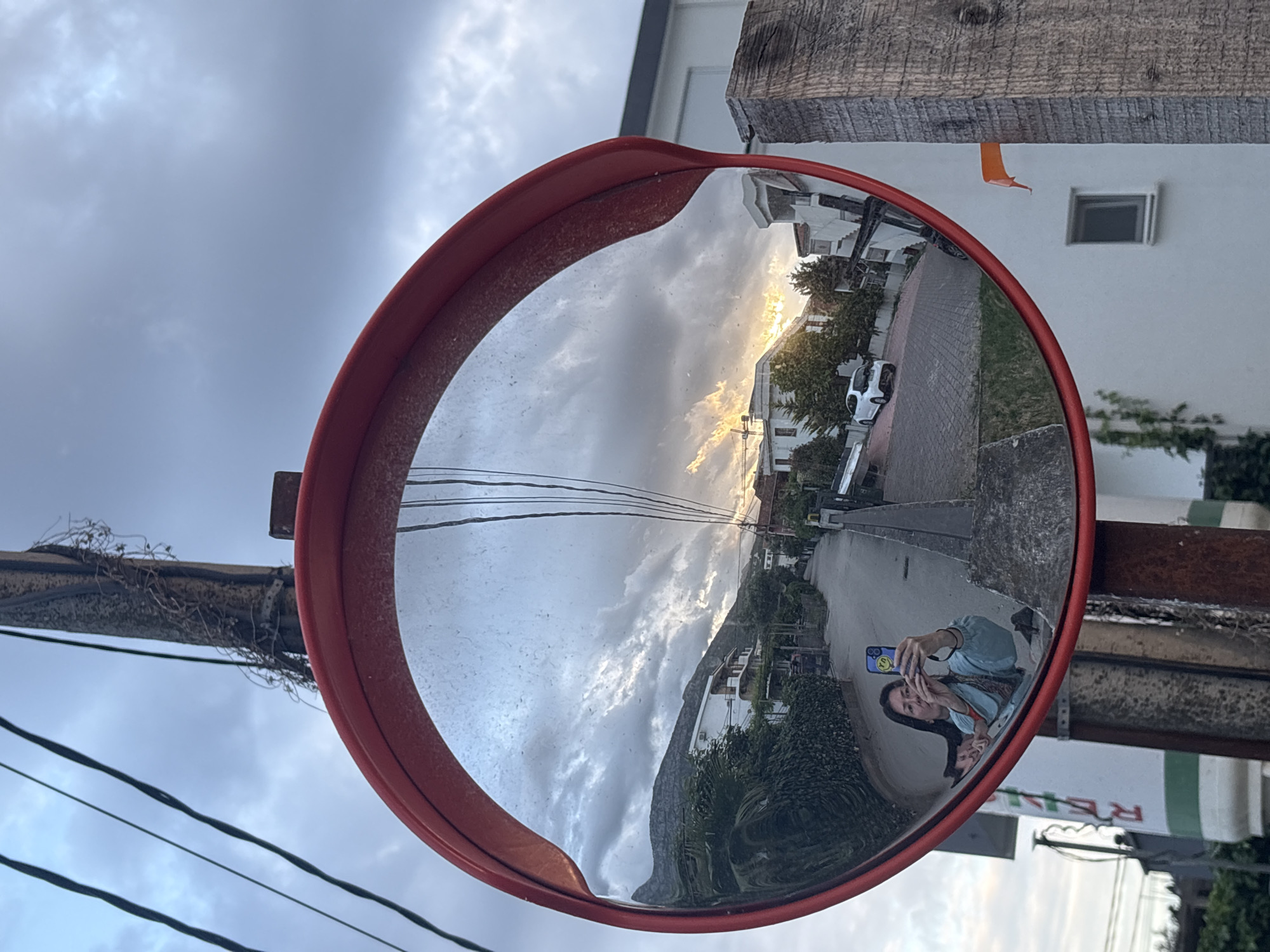 The image depicts a convex mirror, likely a traffic mirror, reflecting a scene in Šušanj, Montenegro. The mirror is mounted on a wooden pole attached to a concrete pole. A woman is visible in the reflection, taking a selfie with her phone. The reflection also shows a street with buildings, trees, and a car. A mountain range is visible in the distance. The sky is cloudy, with some sunlight breaking through. Power lines are visible above the mirror. A building with a window is also visible. A sign for a restaurant is visible behind the convex mirror.