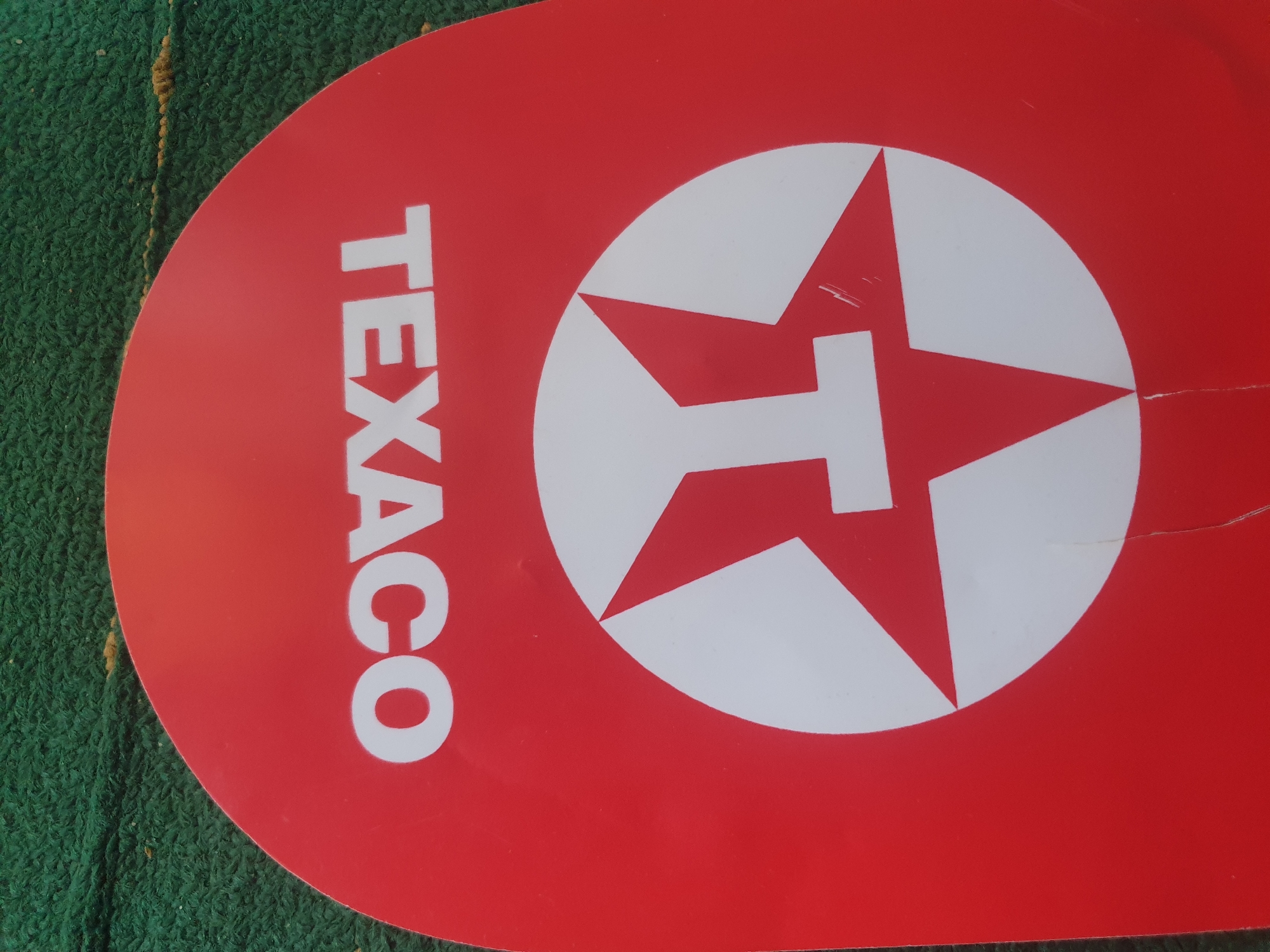 The image displays a red, semi-circular object featuring the white, uppercase text "TEXACO" positioned upside down, centered on its upper half. Below the text, a white circular logo with a red five-pointed star containing a stylized white letter "T" is also visible, oriented upside down. The red object exhibits minor surface scratches, particularly noticeable near the logo's upper left point and along its bottom edge, where a faint vertical crease is also present. This red object rests on a textured, shaggy green surface, possibly a carpet or artificial turf, which shows scattered light brown specks. The overall scene is static, with no people or active interactions depicted. The location is unknown.