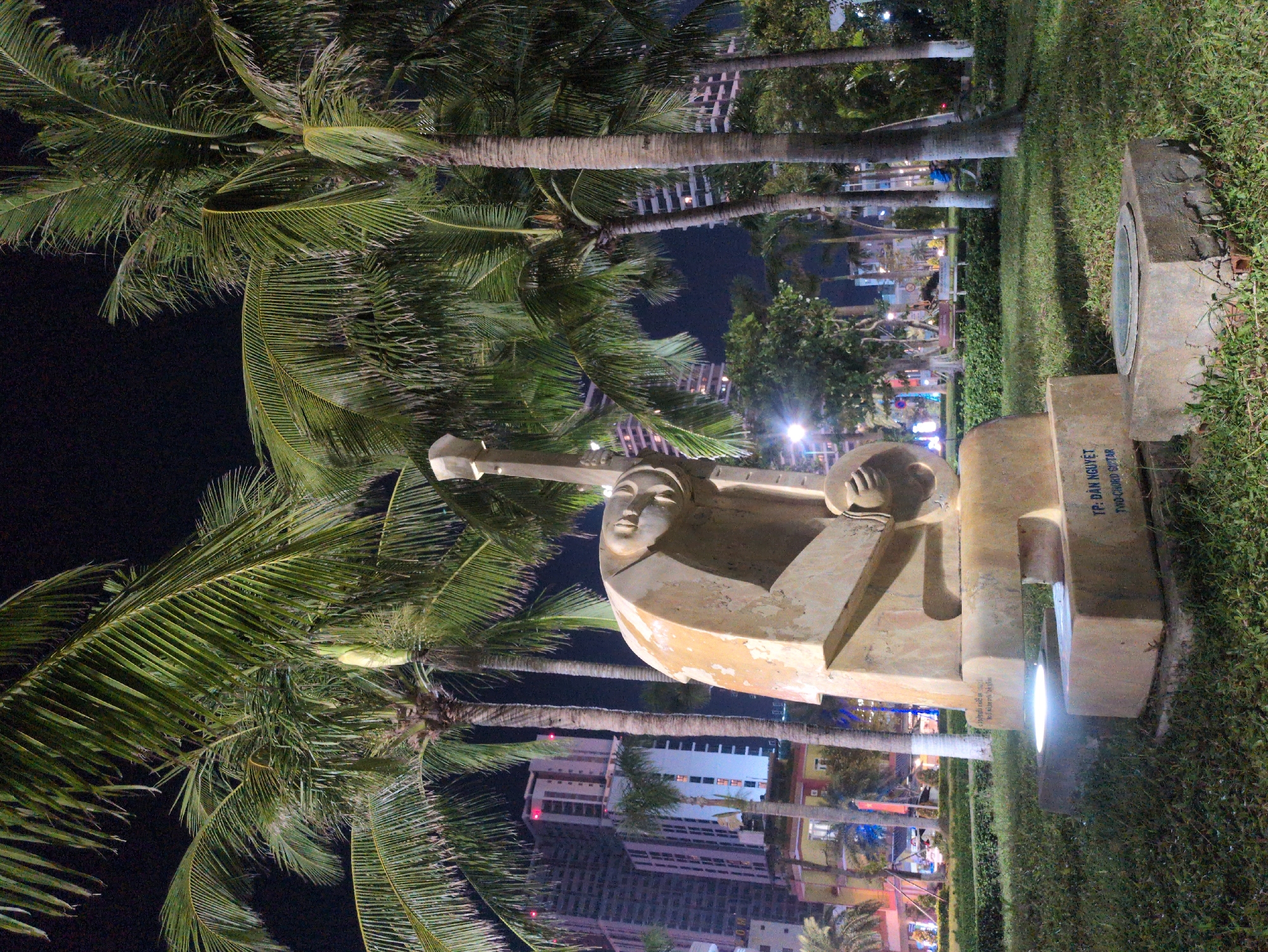 A sculpture of a person holding a guitar stands in a park in South Quy Nhon Ward, Vietnam, at night. Palm trees frame the sculpture and stretch towards the city skyline in the background. The sculpture is made of light-colored material and is illuminated from below. The surrounding area is filled with green grass and other vegetation. The city lights are visible in the distance.