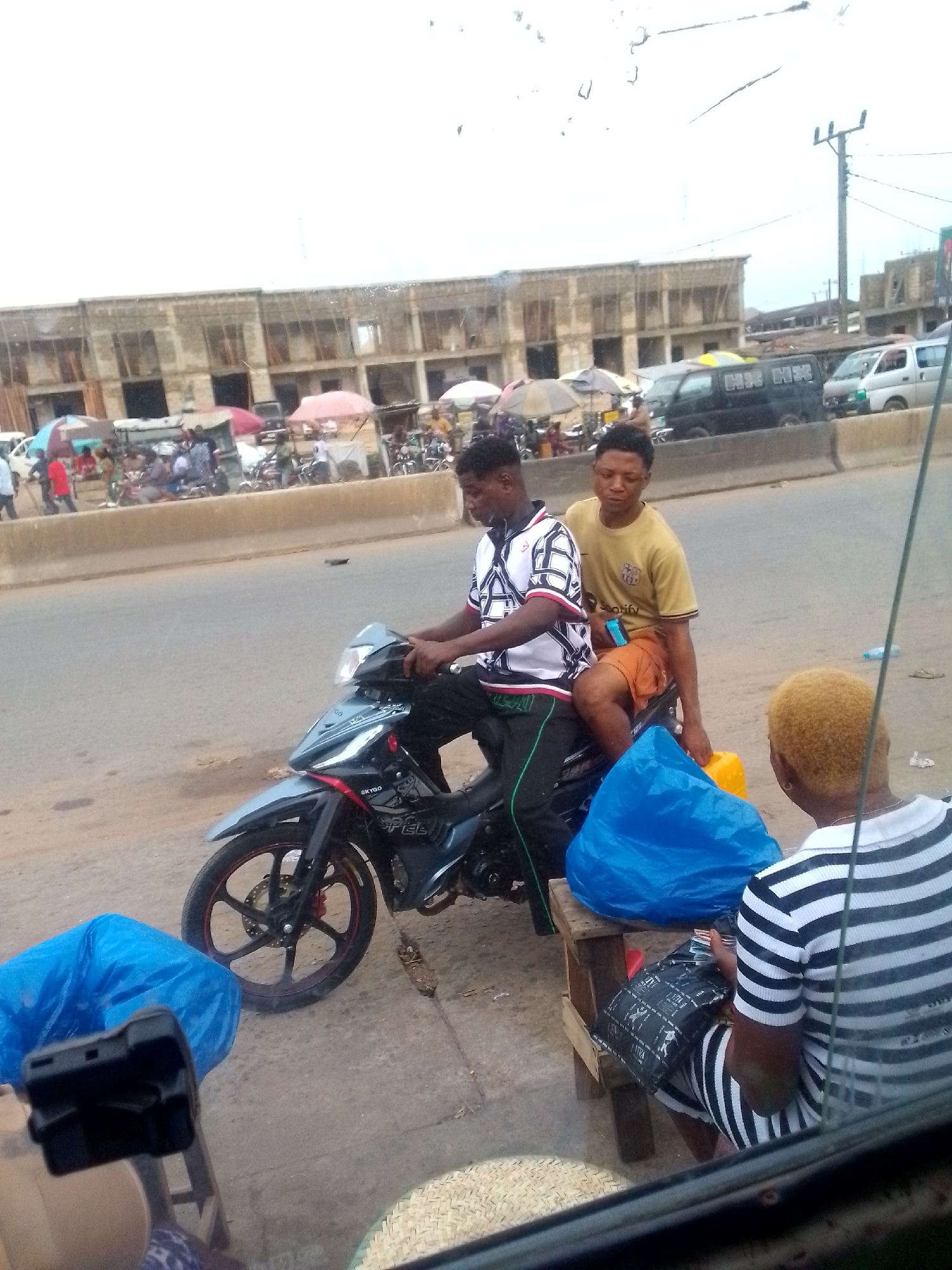 Two men on a black Skygo motorcycle are parked on the side of a road in Ihiala, Nigeria. The driver wears a white shirt with a black-and-white pattern, and black pants with a green stripe. The passenger behind him wears a tan shirt and orange shorts, holding a yellow jerrycan. Behind them, a person in a striped shirt and shorts is seated on a wooden stool, looking at a dark package. Next to them is a blue bag. The background shows a busy street with several vehicles, people, and commercial stalls. A large building under construction is visible in the distance. Overhead power lines are also present.