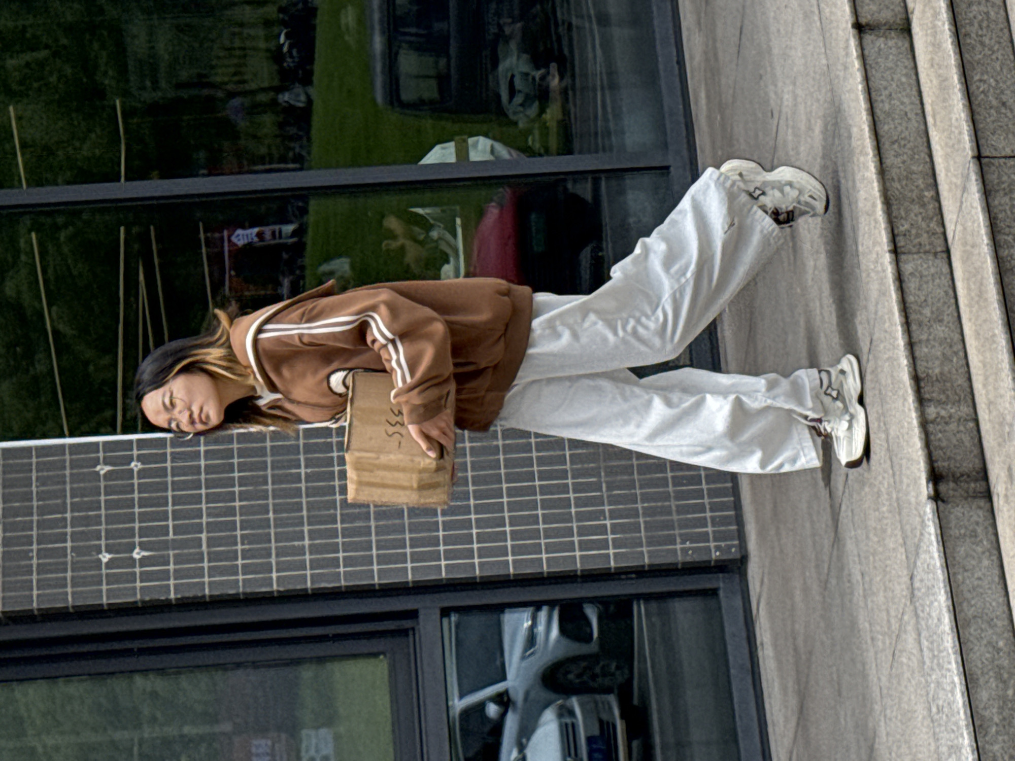 The image, set in Jiangjin, China, shows a young woman walking or standing outdoors beside a modern building during the daytime. She has long, dark hair with lighter streaks and is looking downwards or to her left.
She is dressed in a casual outfit, featuring a brown hooded sweatshirt with white stripes on the sleeves and along the zipper, paired with loose-fitting white pants and white sneakers. She is holding a small, plain cardboard box against her left hip. Partially visible handwritten text on the box includes "335-".
The background consists of a building with large, dark reflective windows that show reflections of green trees and a light-colored car. Below the windows, there's a dark grey tiled wall with a grid pattern. To the right of the woman, a concrete structure with distinct lines, possibly steps or a sloped surface, is visible. The scene is brightly lit by natural light, suggesting clear weather. Her expression is neutral.