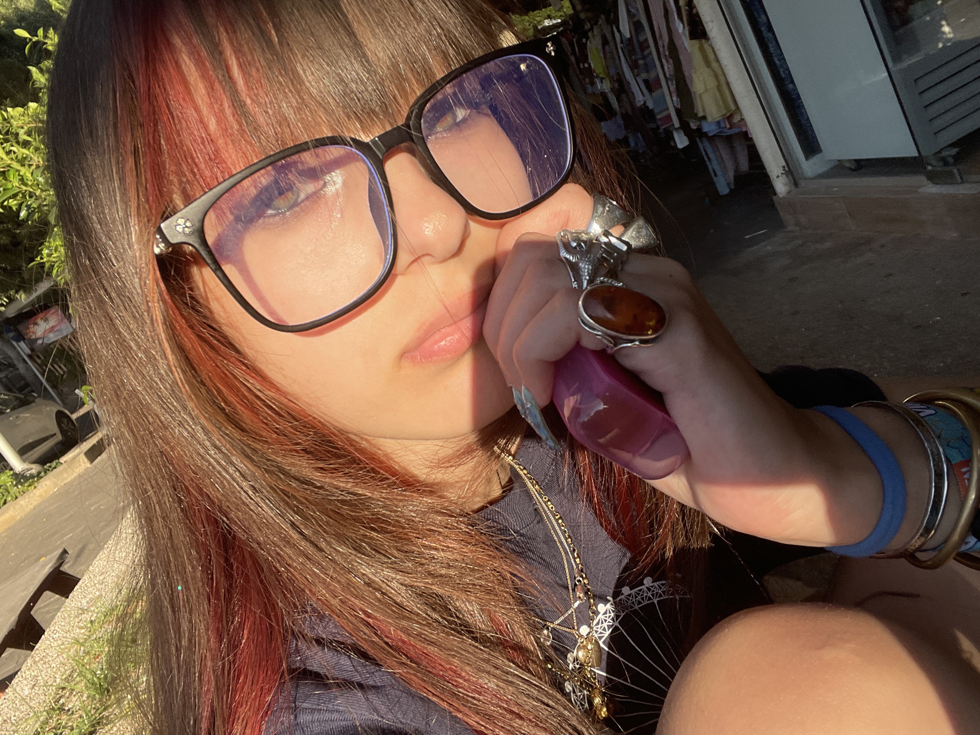 The image is a close-up selfie taken outdoors, likely in Raanana, Israel, during daylight hours, with strong sunlight casting shadows. A young person with brown hair streaked with red is wearing glasses and a dark navy top. Their hand, adorned with multiple rings including one with an amber stone and another with a ram's head design, is holding a pink object. Several necklaces are visible on their chest, layered over the top. The background shows a textured surface that could be a pathway or patio, with some greenery and what appears to be a stack of books or files near a white structure, possibly an outdoor cabinet or shelf. The overall impression is a casual, sunlit moment captured outdoors.