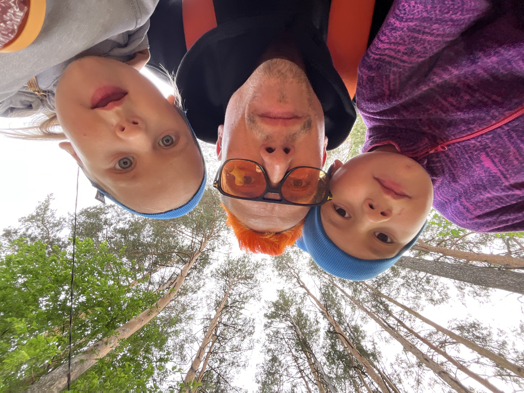 A low-angle shot looking upward depicts three individuals, possibly a parent and two children, standing in a forest. The person in the center has orange hair and is wearing sunglasses. The children on either side of the central figure wear blue hats. The background shows tall trees with green leaves. The location is inferred to be in a forest area in or near the city of Chaikovsky, Russia.