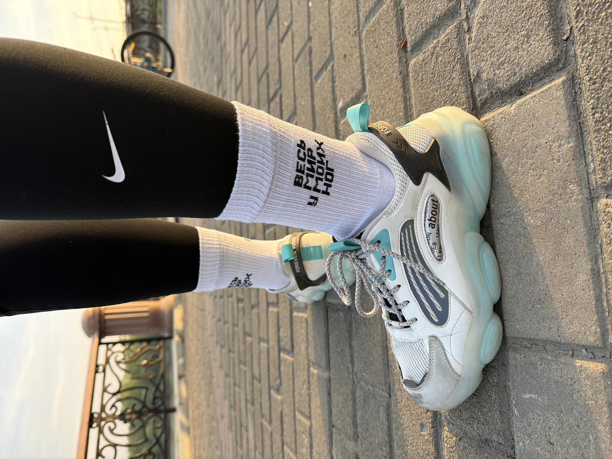 A close-up shot displays a person's lower legs and feet, revealing Nike leggings and white socks with Russian text paired with chunky white sneakers featuring light blue accents. The person stands on a paved walkway composed of rectangular gray stones, likely in an urban setting. A wrought iron fence is seen in the background. The location is possibly in the city of Birobidzhan, Russia.