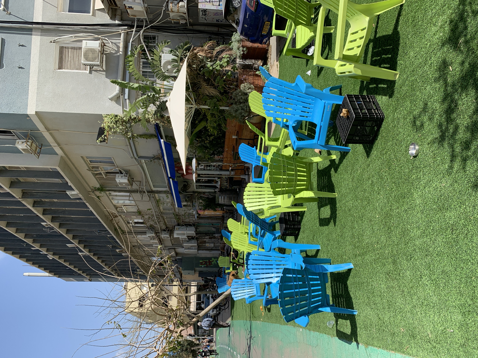 The scene is outdoors on a sunny day in Tel Aviv, Israel. The primary focus is on an area set up with blue and green plastic Adirondack chairs placed on artificial green turf. The seating area appears to be part of a cafe or outdoor space along a pedestrian walkway. In the background is a multi-story building with air conditioning units visible on the exterior. A white umbrella is positioned amidst greenery, providing shade. People are seen walking on the path in the distance, and there are parked bicycles nearby. A dark-colored car is also partly visible in the background. A black plastic crate is located near the chairs.