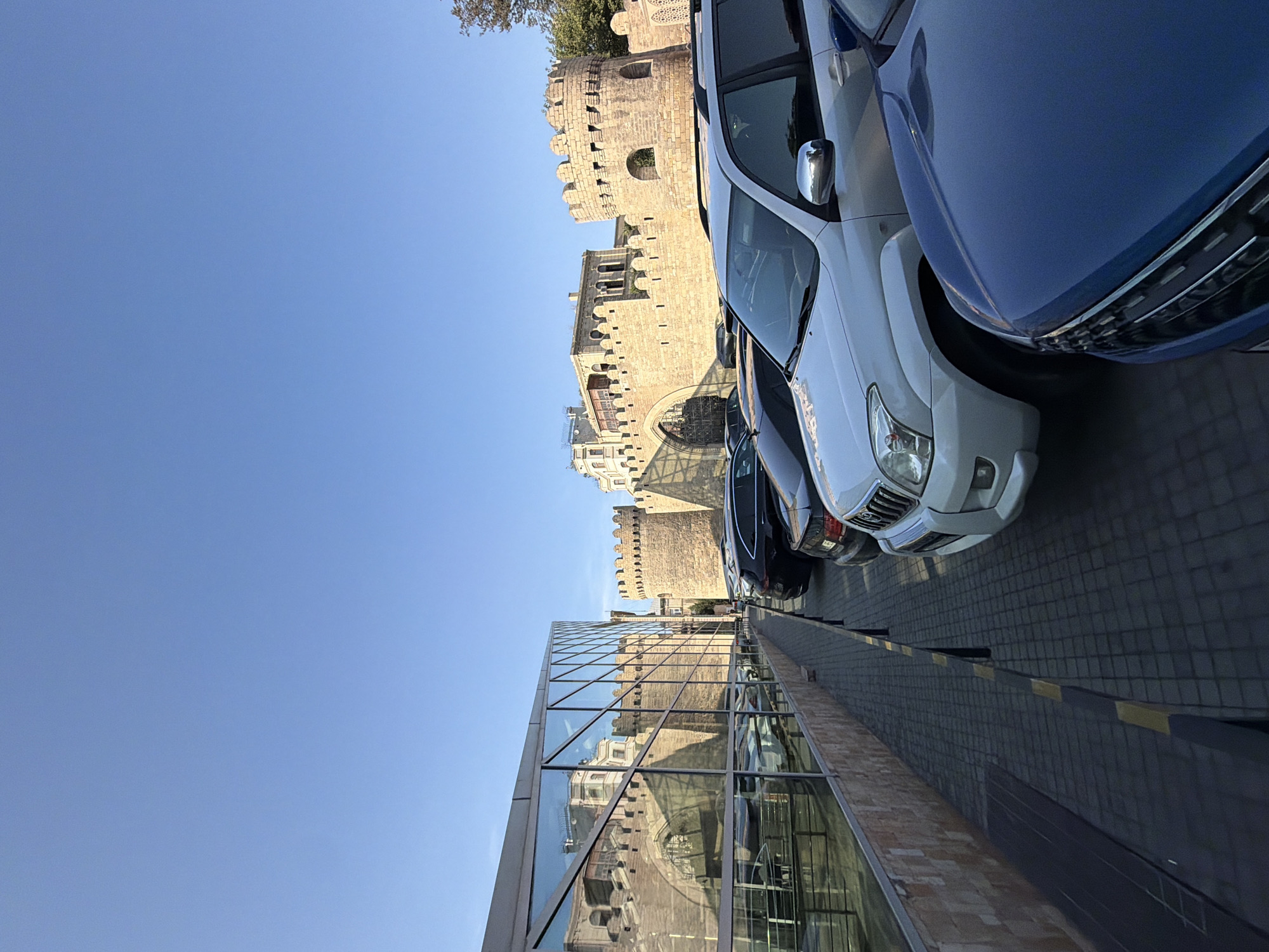 The image depicts a modern building with a glass facade reflecting an ancient stone fortress wall with crenellated battlements and an arched gateway. The fortress appears to be in the Sabail Raion district of Azerbaijan. In the foreground, several parked cars are visible, including a white SUV and a blue car. The cars are parked on a paved area adjacent to a sidewalk. The sky is clear and blue, indicating daytime.