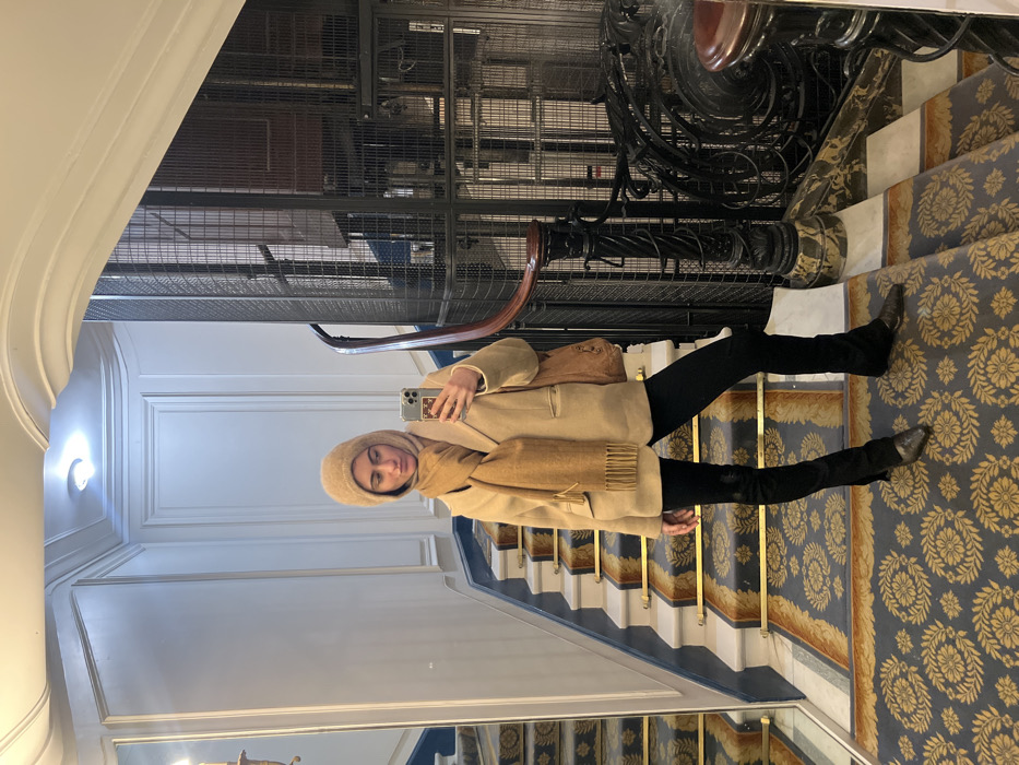 A woman stands on a patterned staircase, taking a selfie. She wears a light brown coat, scarf, and hat, paired with black leggings and boots. An elevator shaft is visible behind her. The staircase features marble accents and a blue and gold floral carpet. The interior suggests a building with historical architectural details.
