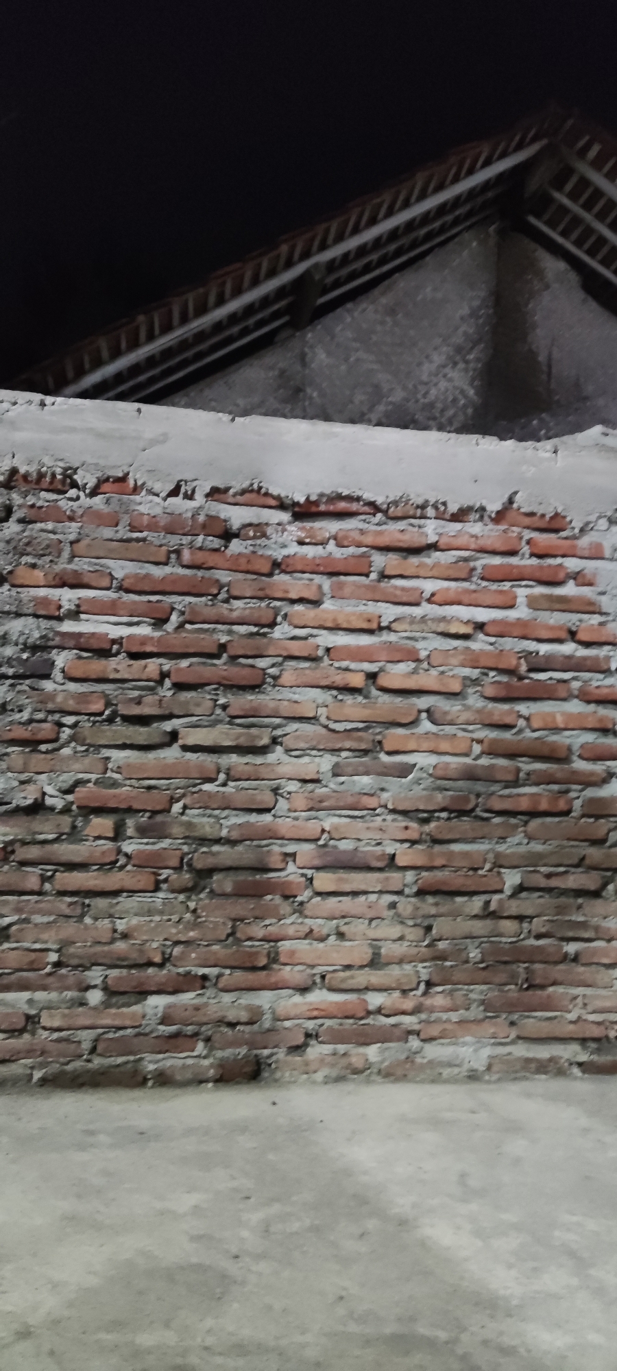An outdoor, low-angle medium shot captures a section of a brick wall and the lower portion of a roof against a dark night sky. The wall is constructed from red bricks, some showing signs of weathering and minor imperfections, with gray mortar filling the gaps between them. Above the brick wall, a rough layer of gray concrete creates an uneven cap. The roof, visible at the top of the frame, has exposed wooden beams and what appear to be terracotta tiles. The ground in the foreground is a concrete surface, appearing aged and possibly dusty. The scene suggests an outdoor, unfinished construction or an older building in a dimly lit environment, likely in Ciledug, Indonesia. No people are present in the image, and no specific actions or interactions are depicted.