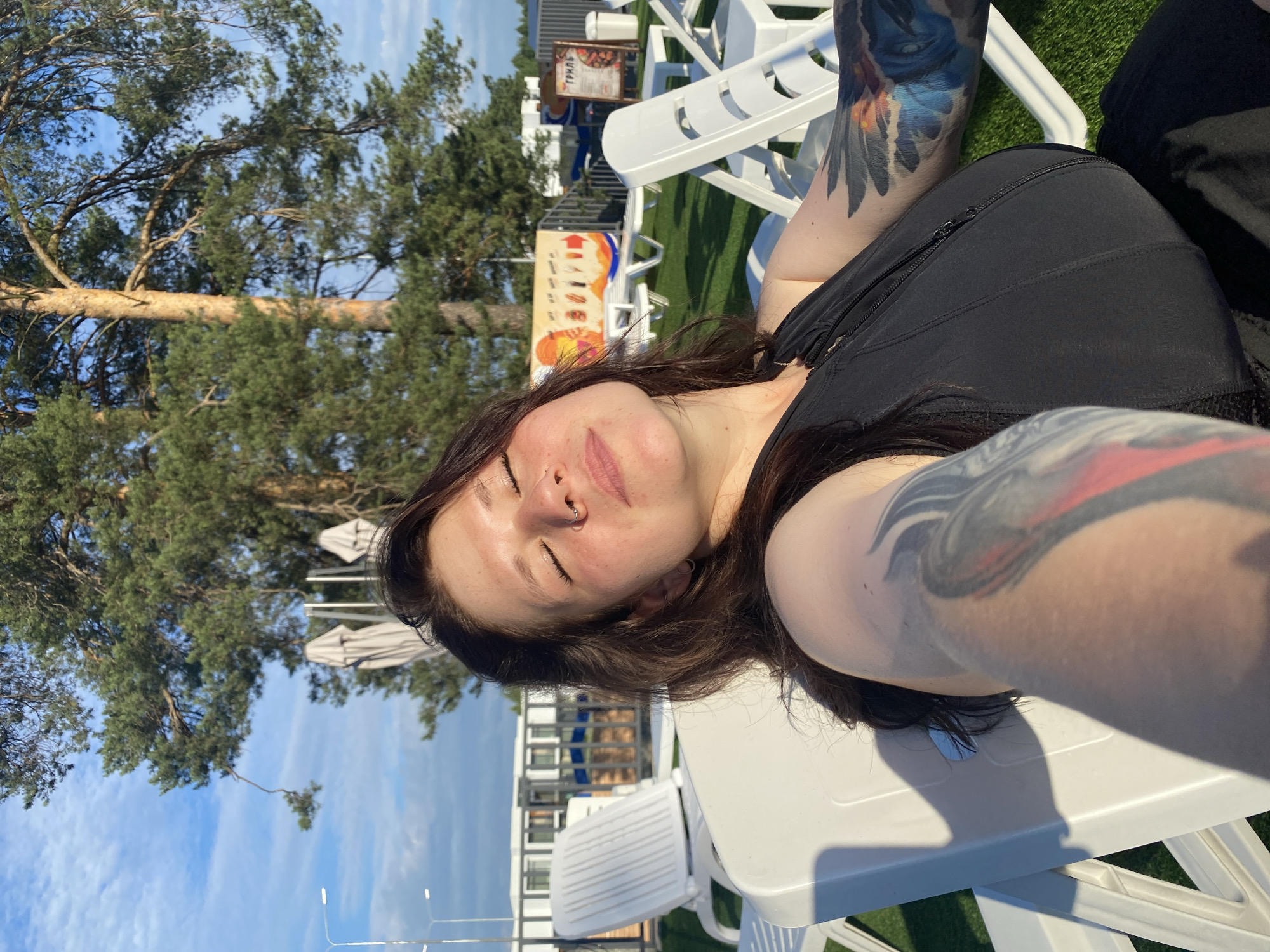 A woman with dark hair and tattoos on her arms is relaxing on a white plastic lounge chair outdoors. She is wearing a black top with a zippered front. Her eyes are closed, and her head is tilted back, appearing to be enjoying the sunlight. In the background, there are more white lounge chairs, a large pine tree, and some buildings with advertisements. The scene is set on a grassy area, possibly at a beach or resort. The presence of pine trees and buildings suggests a location within or near a park or recreational area, potentially within the city of Votkinsk, Russia, given its natural surroundings.