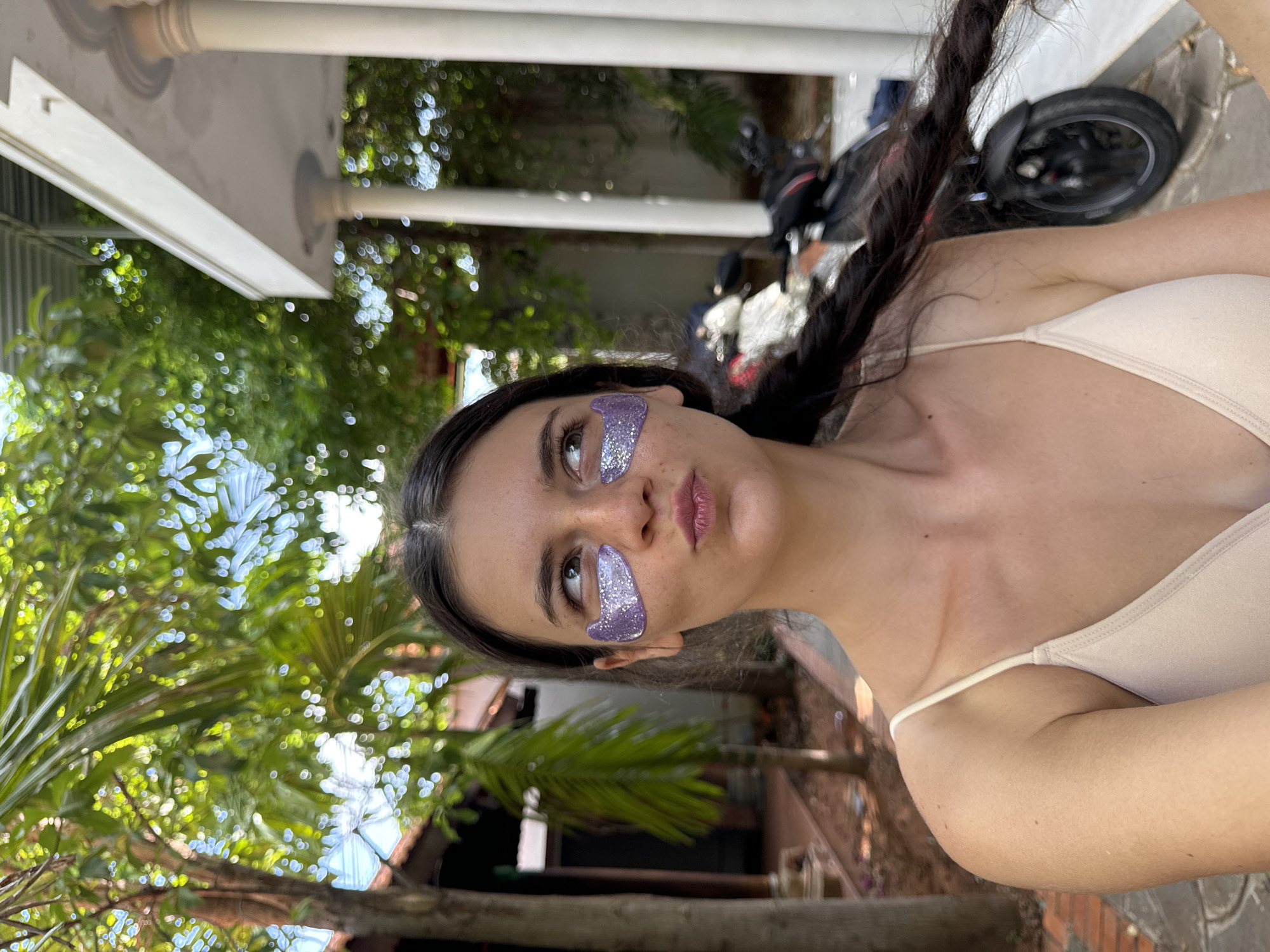 A young woman with dark hair in a braid is positioned in the foreground of an outdoor scene. She is wearing a light-colored tank top and has purple, glittery under-eye patches. Her mouth is pursed, and she is looking to her right. Behind her, there is lush green foliage, including palm trees, and parts of a building with white columns and a light-colored roof. A motorcycle is visible in the background. The overall setting appears to be a residential or garden area.